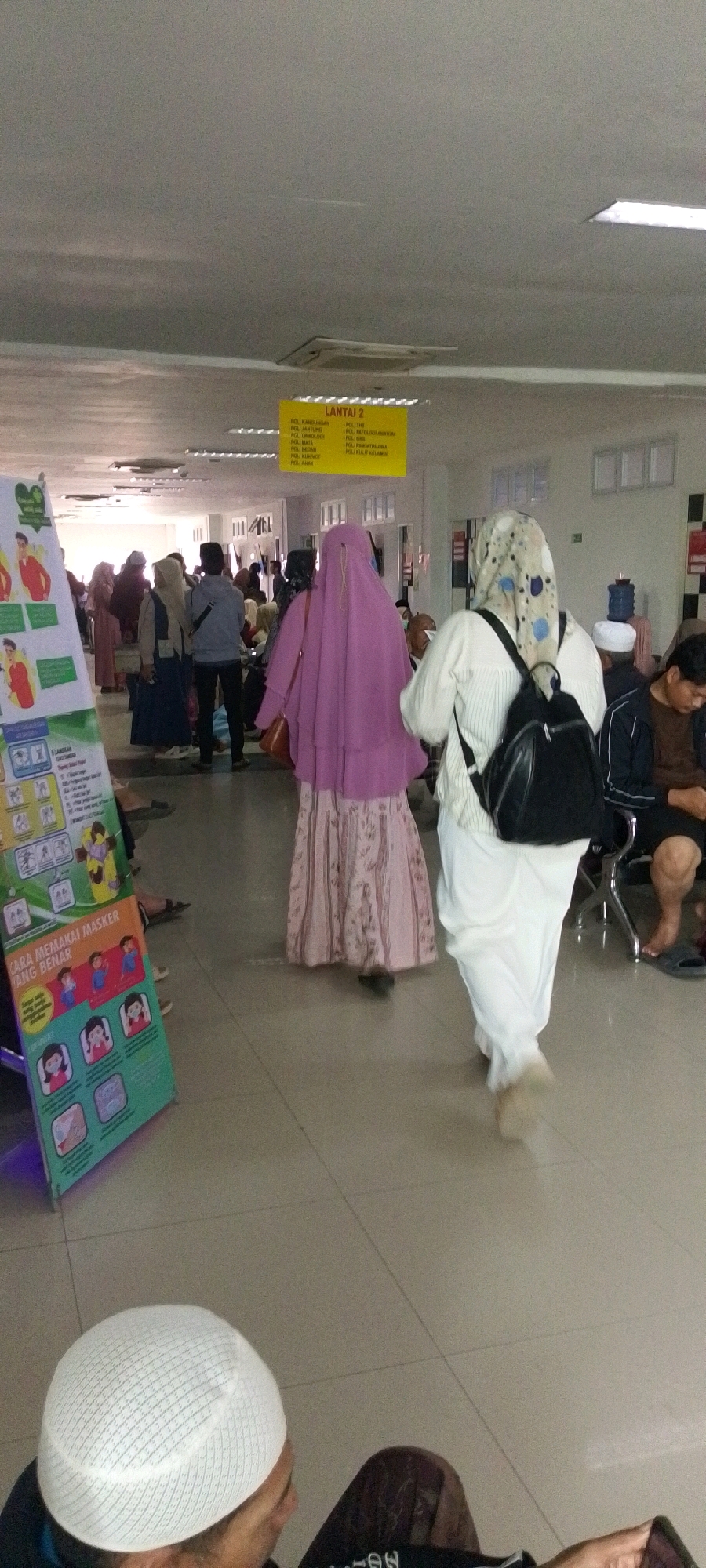 The image depicts an indoor scene, likely a public waiting area, with a tiled floor and a dropped ceiling. Several people are present, some walking and others seated. The individuals are predominantly wearing modest clothing, with many women in hijabs. In the foreground, a person wearing a white skullcap is visible from the shoulders up, looking down.
Further into the scene, a woman in a long, pale pink dress and a light purple hijab is walking away from the camera. To her right, another woman in a white outfit and a patterned headscarf with a black backpack is also walking away. A green and white informational poster is positioned on the left side of the frame, displaying illustrations and text in Indonesian, including the phrase "CARA MEMAKAI MASKER YANG BENAR" (How to Wear a Mask Correctly).
Above the people, a yellow sign is suspended, indicating "LANTAI 2" (Floor 2) and listing various medical departments or clinics. The background shows a hallway with doorways and what appear to be chairs and other people waiting. The overall impression is of a busy, functional space, possibly a hospital or a government building with medical services. The presence of the mask poster suggests the photo was taken during a period where mask-wearing was emphasized. There is no specific visual information in the image that would allow for the precise identification of the city of Sumenep, Indonesia, beyond it being a general indoor public space.