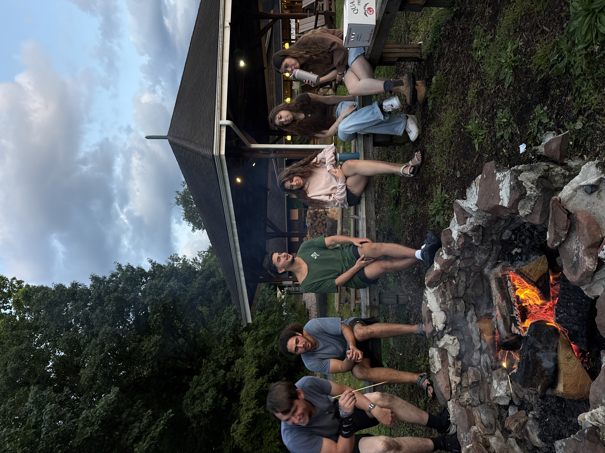 An outdoor evening scene depicts six young adults gathered around a stone fire pit. In the foreground, a low, irregularly shaped fire pit contains burning logs and glowing embers, emitting smoke. A light-skinned male with short dark hair, wearing a grey t-shirt and dark shorts, leans forward on the left, holding a long stick towards the fire. To his right, another light-skinned male with long curly dark hair, wearing a blue t-shirt and dark shorts, sits with clasped hands, observing the fire. Further right, a third light-skinned male with short dark hair, dressed in a dark green t-shirt and dark shorts, sits facing right, away from the fire.
On the opposite side of the fire pit, three light-skinned females are seated on a wooden bench. The leftmost female of this group, with long wavy dark hair, wears a light pink hoodie and dark shorts, looking downwards. A blue thermal bottle rests beside her. Next to her, a female with long wavy dark hair, clad in a dark t-shirt and light blue jeans, looks forward. The rightmost female, with long wavy brown hair, wears a brown long-sleeved shirt and light blue denim shorts, and is actively drinking from a large silver thermal bottle. A white cardboard box, partially visible with text "QUA..." and "GROWN & PACKED IN U.S.A.", sits on the bench beside her.
All individuals are seated on rustic wooden benches arranged around the fire pit. In the mid-ground, a large, dark-roofed pavilion or gazebo with white trim and support columns is visible. String lights illuminate the underside of its roof, indicating the evening hour. A stack of firewood is present near the pavilion's base. Dense, dark green deciduous trees frame the left side and background of the scene. The sky above is cloudy, displaying a mix of grey and light blue patches. The ground is a mix of dirt, sparse grass, and mulch. The setting is consistent with a recreational area in Washington Township, United States of America.