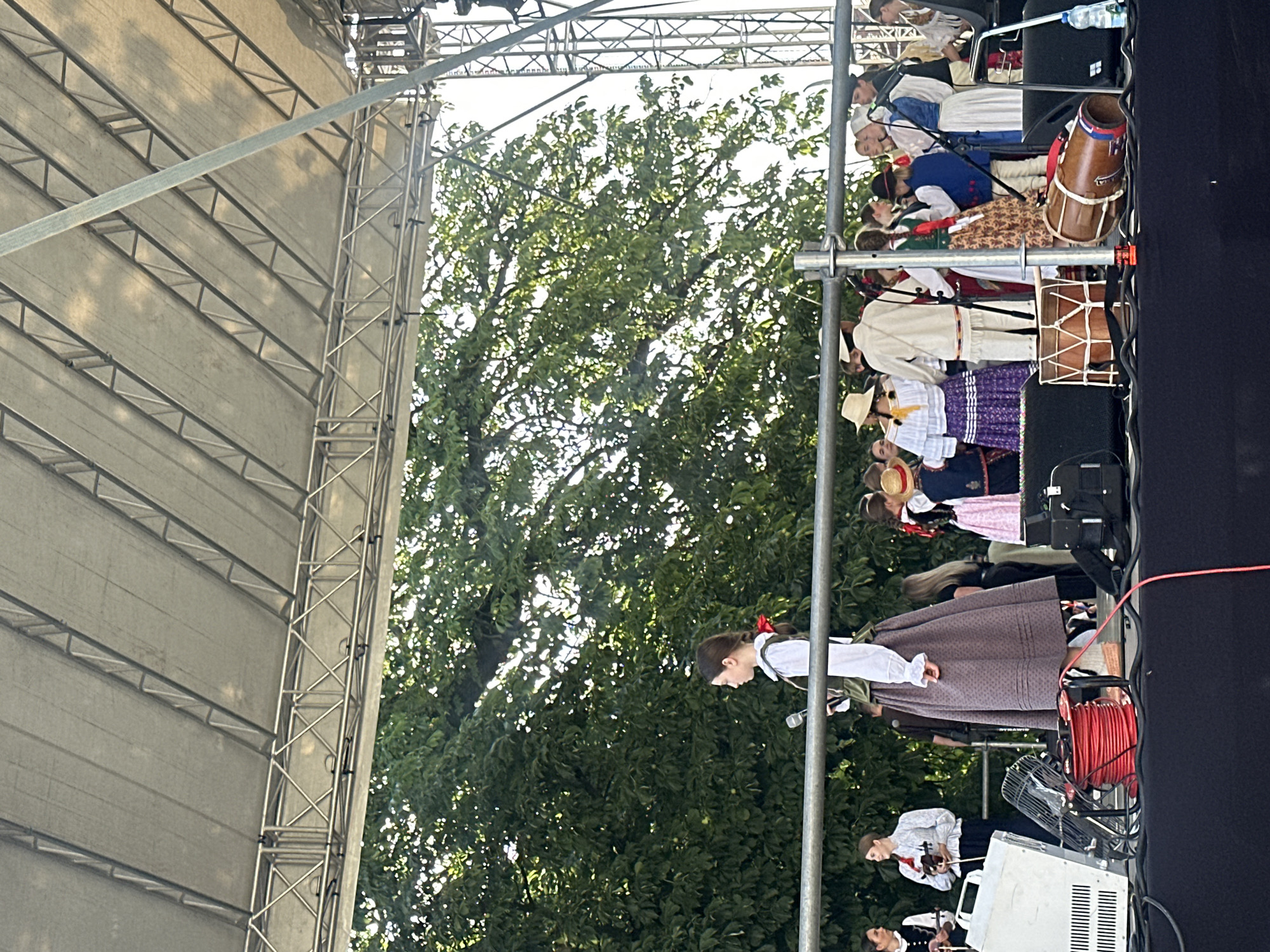 The image depicts an outdoor performance on a stage, likely in the city of Nowy Sącz, Poland. The stage is covered by a large, tent-like structure supported by metal scaffolding. Behind the stage, a dense canopy of green trees is visible against a bright sky.
On the stage, a group of people are gathered, many dressed in traditional folk costumes. In the foreground, a woman in a dark skirt and white blouse with a red ribbon in her hair holds a microphone. To her left, two individuals are visible, one playing a violin. In the background, other performers are dressed in various colorful traditional outfits.
Several musical instruments are placed on the stage, including two drums, one larger and one smaller, positioned in the lower right and lower middle sections of the image respectively. Electrical equipment, including a white speaker or amplifier and a black case, are also present on the stage floor, along with coiled red cables and a black fan. A water bottle sits on a speaker in the far right. The overall scene suggests a cultural event or folk music performance.