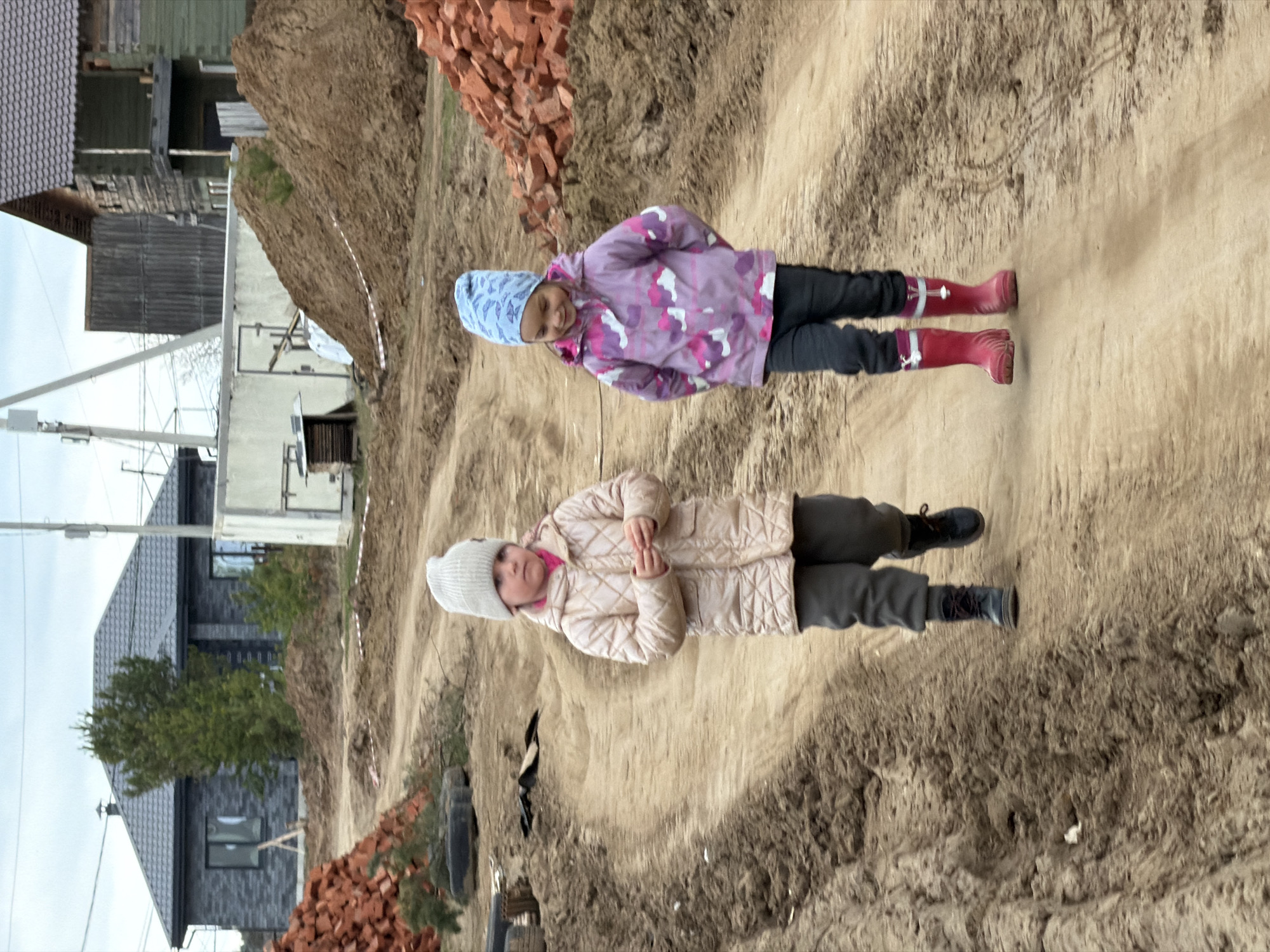 The image depicts two young girls standing on a dirt path near a construction site in what is likely the city of Chaikovsky, Russia.
The girl in the foreground wears a light-colored quilted coat, gray pants, black boots, and a white knitted hat. She has her hands clasped in front of her.
The girl behind her is wearing a purple camouflage jacket, black pants, red boots, and a light blue knitted hat. Her hands are also clasped in front of her.
The path is dirt or sand. To the left are houses, some under construction, and stacks of red bricks. The sky is overcast.