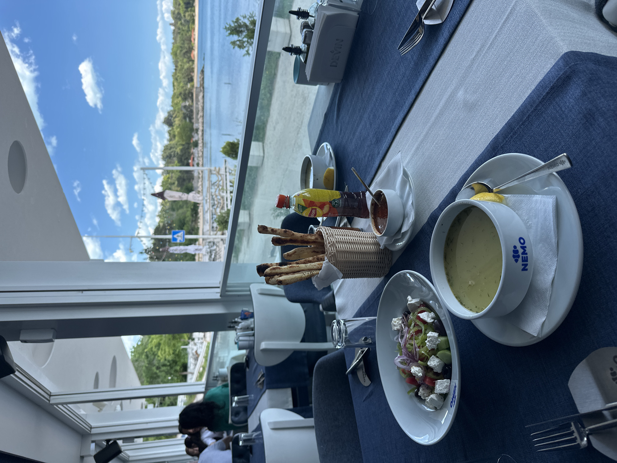 The image depicts a table setting at a restaurant in Varna, Bulgaria. The view from the restaurant shows the sea and part of the city, likely near the Primorski Park.
On the table, which has a blue and white striped runner, are several food items:
- A bowl of soup with the "Nemo" logo on the side, placed on a saucer with a lemon wedge and a napkin.
- A salad with feta cheese, tomatoes, olives, and red onions in a white oval bowl.
- A basket of breadsticks.
- A bottle of juice and a small bowl of a dark reddish sauce, possibly ajvar or a similar pepper-based spread.
- A cup with a brown beverage on a saucer, next to the condiments.
Cutlery is placed at the edge of the table.
Part of the restaurant's interior is visible, including chairs and other tables. People are seated at the table nearby, visible behind the back of the chairs.
