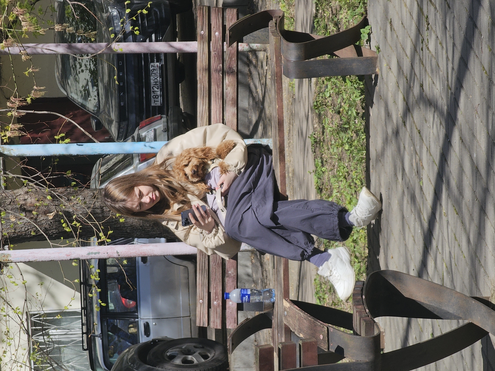 A woman sits on a park bench, holding a small brown dog and looking at a smartphone. The bench appears to be in a public area in the city of Khimki, Russia, near parked cars with Russian license plates (T812HX797). A bottle of water sits on the bench near the woman. The area has trees and paved walkways. The woman is wearing a beige jacket, dark blue trousers, and white sneakers.