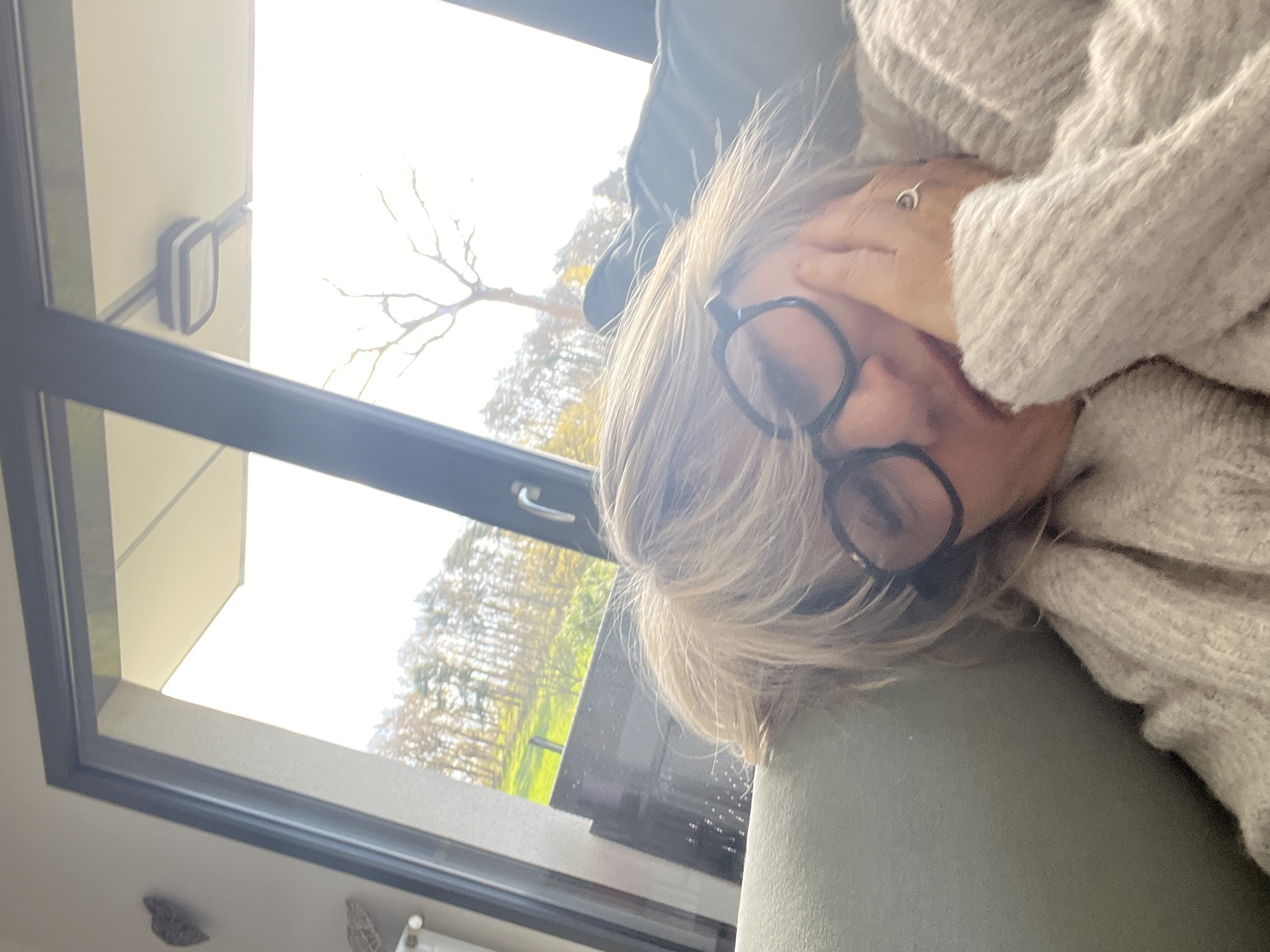 The photograph captures a fair-skinned person lying down, possibly on a sofa, with a light-colored knitted sweater partially covering their face. The person is wearing black-rimmed glasses. One hand is raised, partially obscuring their face. The location appears to be indoors, next to a large window. Outside the window, trees and a field are visible, suggesting a natural or semi-rural setting. There are no other people visible in the image. The photo was taken in Chambœuf, France.