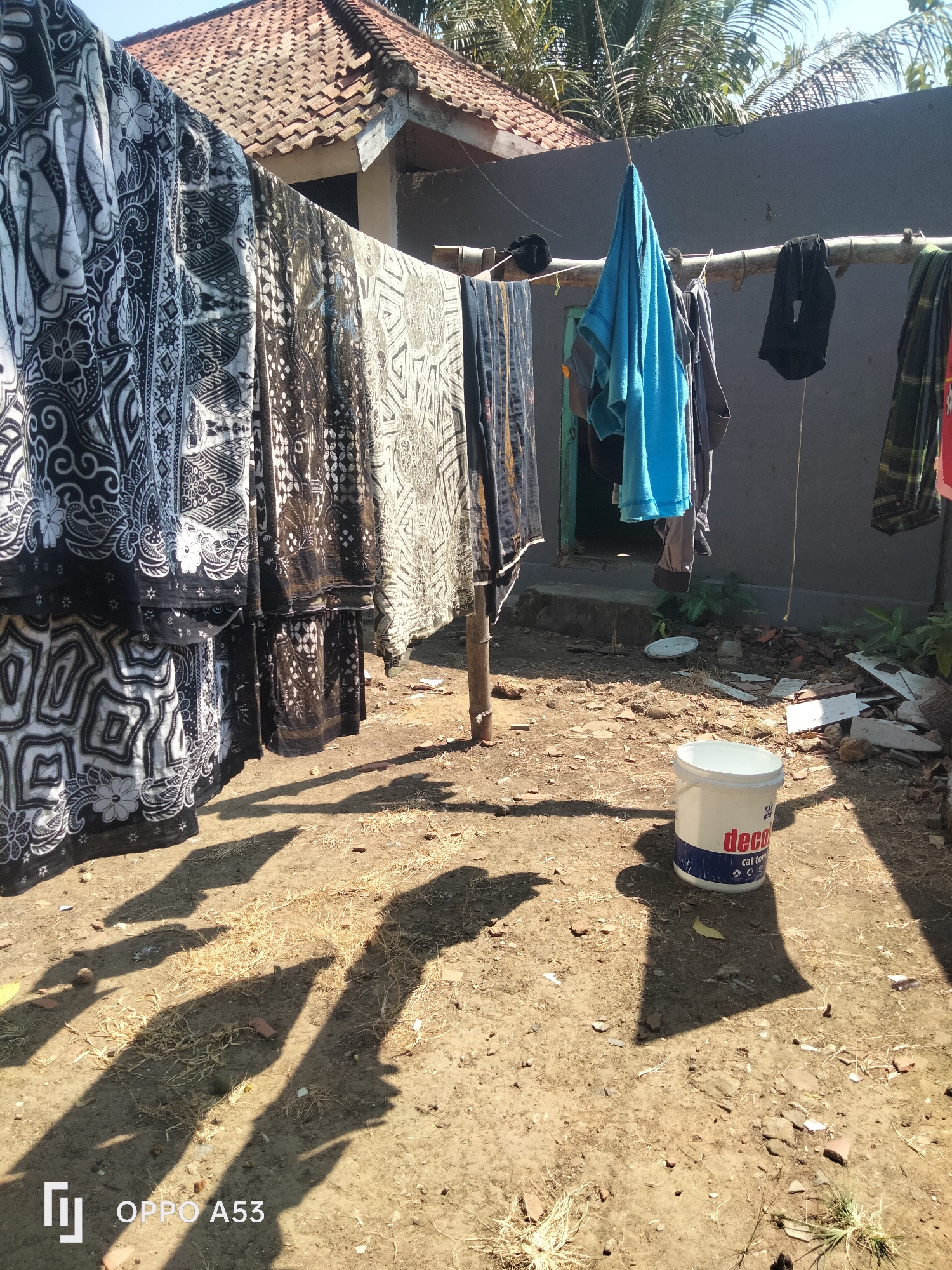 This is a daytime, eye-level, outdoor shot of laundry drying on a clothesline in what appears to be a backyard in Badung, Indonesia. The clothesline is made of bamboo poles, and various items of clothing are hung, including several pieces of batik fabric with intricate black and white patterns, a bright blue towel, and dark-colored garments. The background shows a simple house with a tiled roof and a gray wall. The ground is dry and dusty, with scattered debris like wood scraps and broken tiles. A white plastic bucket with red and blue lettering, possibly a paint can, sits on the ground in the foreground, casting a long shadow. The scene is illuminated by bright sunlight, creating distinct shadows on the ground.