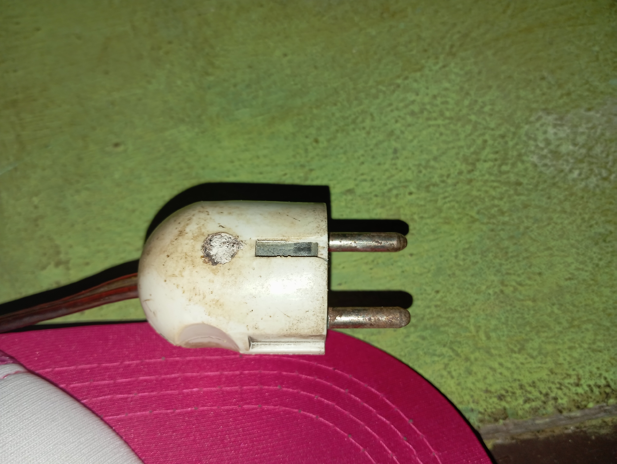 A close-up, waist-level outdoor shot captures a weathered, white electrical plug against a textured, greenish-yellow wall. The plug is partially obscured on the left by a bright pink, fabric object, possibly a hat or bag. Two metallic prongs extend downwards from the plug. The wall's surface appears rough and mottled, indicating an aged or outdoor environment. The lighting suggests it is daytime. No people are visible. The scene is located in Rejosari, Indonesia.