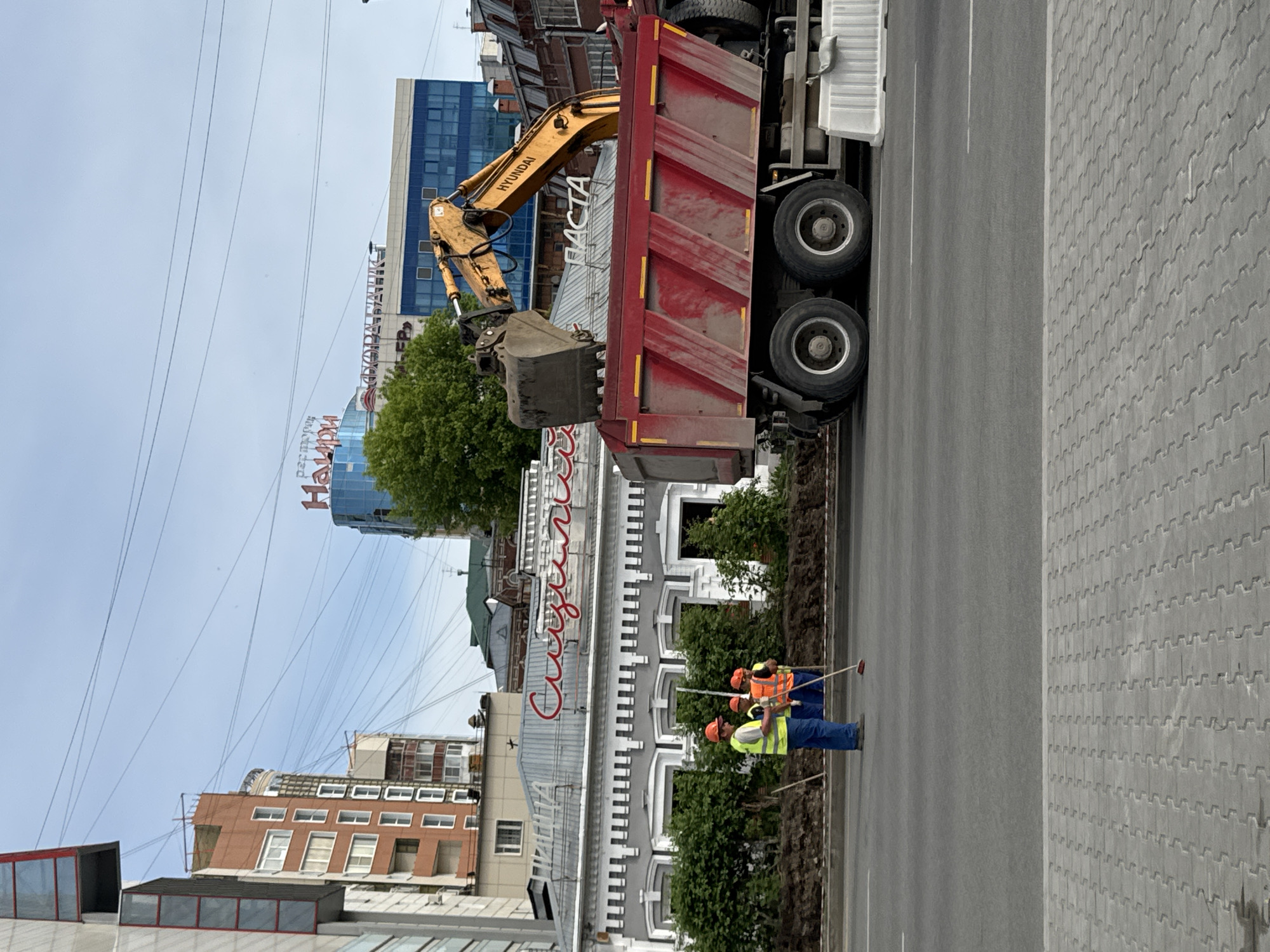 The scene captures urban roadwork in Perm, Russia, along a street lined with buildings.
Dominating the foreground is a red dump truck with a yellow Hyundai excavator positioned to load it. Three construction workers, dressed in blue work pants and high-visibility vests (one yellow and two orange), use brooms near a newly planted strip of greenery dividing the road from the sidewalk, suggesting cleanup efforts.
In the background, the urban architecture includes older buildings, alongside more modern structures with glass facades and signage in Russian. Overhead, a network of utility lines crisscrosses the sky.
