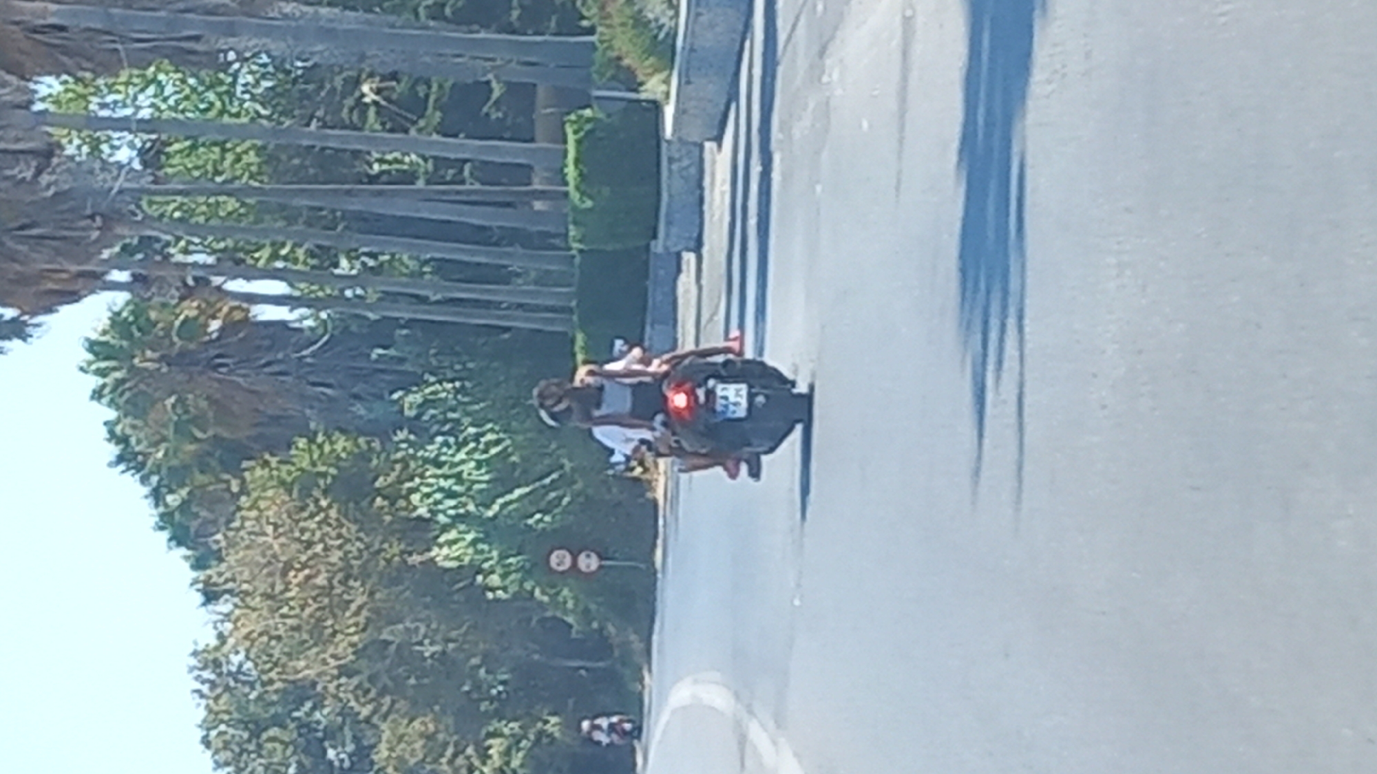 Two individuals are riding on a black scooter on a paved road. The person in front is wearing a white t-shirt and the person behind is wearing a gray top. Both have dark hair and appear to be wearing helmets. The scooter has a red taillight and a license plate that is partially visible. The road is bordered by a grassy median with some bushes and palm trees. In the background, there are more palm trees and foliage, suggesting a warm climate. There are also two circular red and white traffic signs on the side of the road. The scene appears to be in a suburban or semi-rural area of Ηρακλής, Greece.