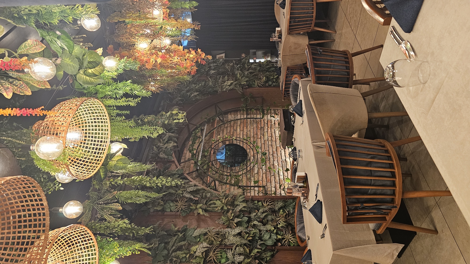 This image depicts the interior of a well-decorated restaurant or cafe, characterized by a lush, botanical aesthetic. The space is adorned with an abundance of artificial greenery, including various types of ferns, leafy plants, and some reddish-orange foliage, cascading from the ceiling and covering wall sections.
Warm, ambient lighting is provided by numerous exposed bulb string lights integrated within the foliage, alongside larger light fixtures featuring natural woven basket shades. One prominent wall boasts exposed red brick, centered by a large, dark circular window or mirror framed by intricate metalwork and winding vines, suggesting an industrial-chic or garden-inspired design. The floor is covered with light-colored tiles.
Multiple tables are visible, uniformly set with light-colored tablecloths. Each place setting includes silverware (fork and knife), an empty glass, and a folded dark napkin. The seating consists of wooden chairs with slatted backs and dark cushions, as well as some chairs with upholstered beige backs. No people are present, but the neatly arranged tables indicate the venue is prepared for diners.
The scene conveys a cozy and inviting atmosphere, suggesting it is evening or nighttime due to the reliance on artificial lighting. The combination of rustic elements like brick and natural materials with abundant greenery creates a vibrant yet intimate ambiance. There is no legible text visible within the image itself. The location context provided indicates this establishment is in Varna, Bulgaria.