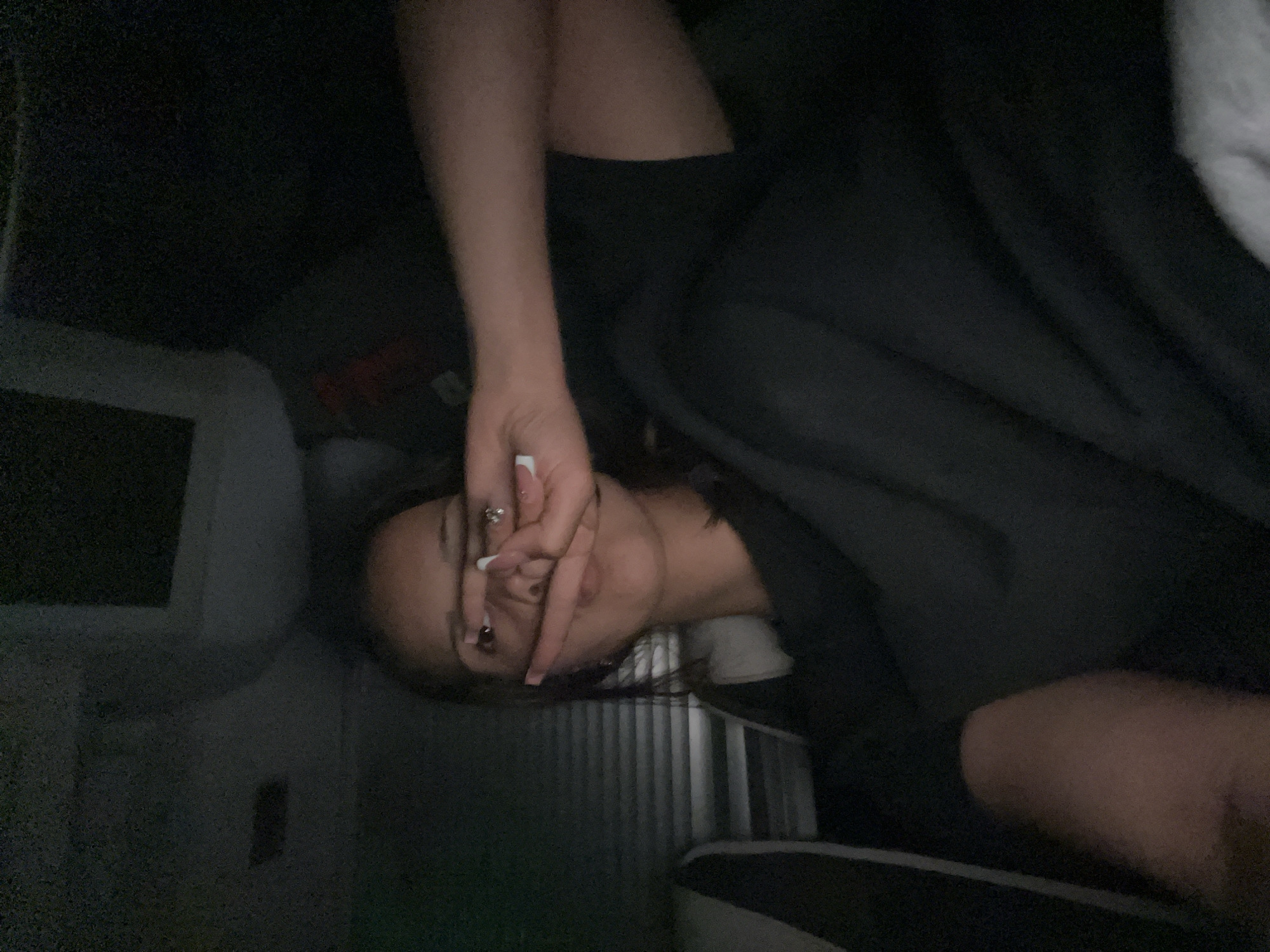 A person is lying down indoors. The person is looking at the camera and their left hand is held up in front of their face. The location is dimly lit. A gray seat is partially visible on the left side, along with a headrest and a screen. The person is wearing glasses. A slatted partition is partially visible behind the person. The person is wearing black clothing and has long dark hair.
