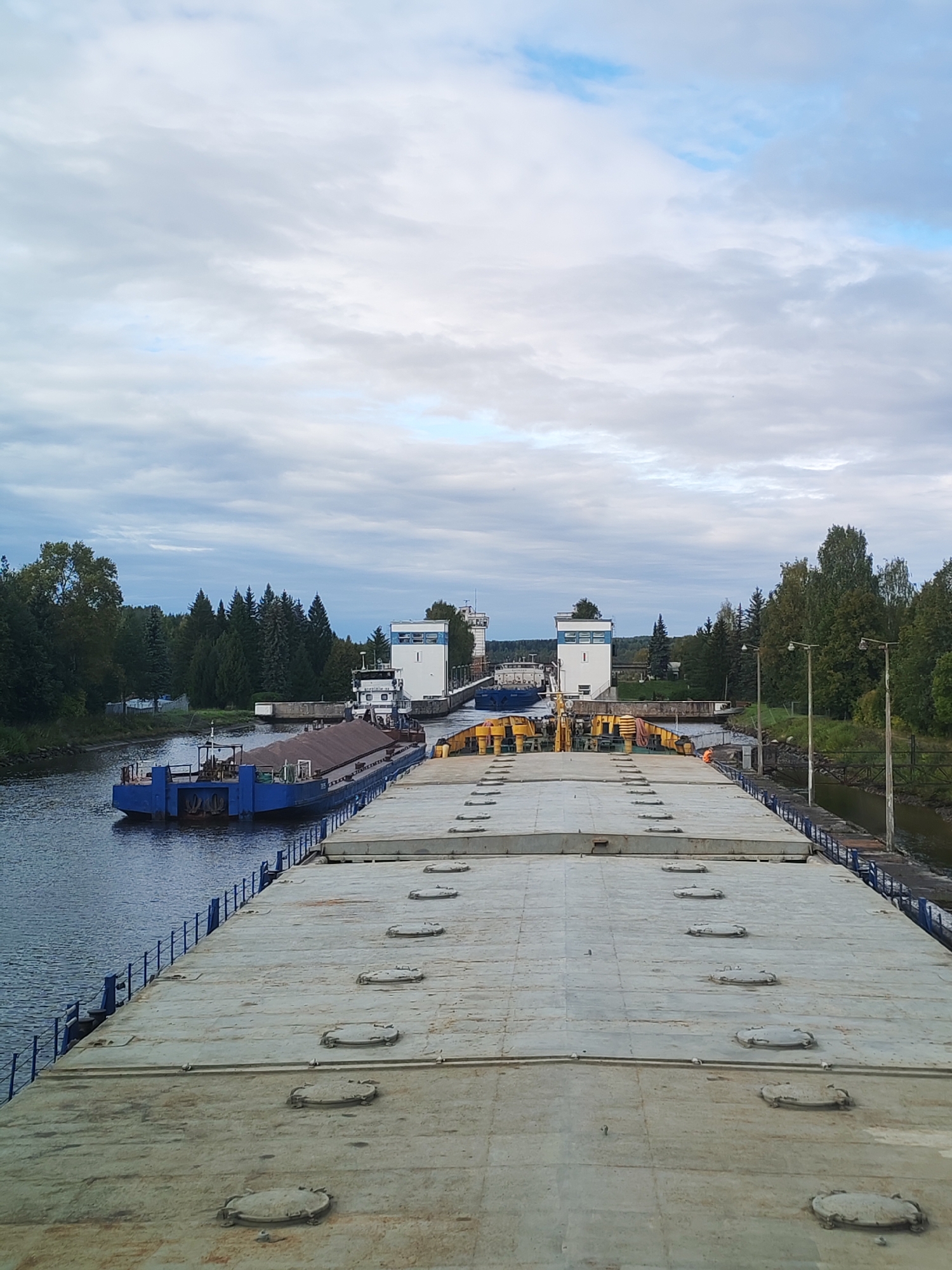 The image shows a view from the deck of a large barge on the Svir River, approaching a lock system in Vytegra, Russia. The barge is laden with a cargo of dark brown material, possibly sand or gravel, covered by a tarpaulin. In the distance, two lock chambers are visible, with a white and blue tugboat docked inside one of them. A blue cargo barge is also visible to the left of the lock. The scene is set against a backdrop of lush green trees and a cloudy sky. The water in the river is dark and calm. The lock structures appear to be modern, with white rectangular buildings and metal gates. The overall impression is one of industrial activity and transportation on a waterway.