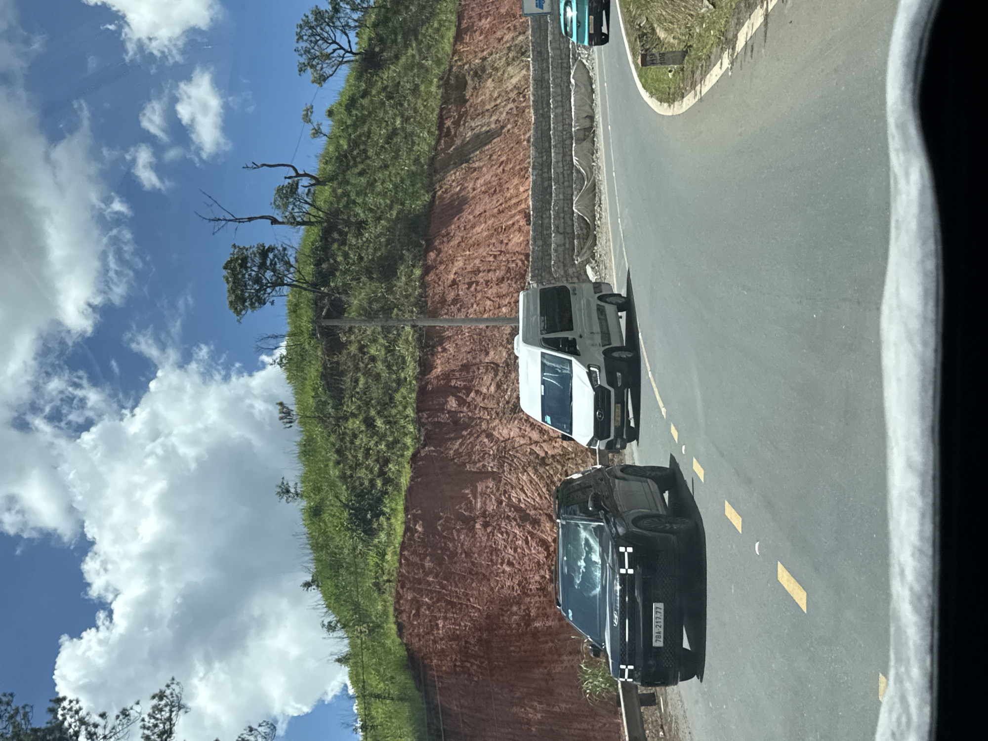 A black SUV with license plate 7BA-217.77 is traveling on a winding road in Thôn 6, Vietnam. A white van is in the lane to the right of the SUV, and a teal car is visible further down the road. The road is bordered by a steep, red-soiled embankment covered in green grass and trees. The sky is blue with scattered white clouds. The scene appears to be a sunny day, likely in the afternoon. The perspective suggests the image was taken from inside another vehicle.