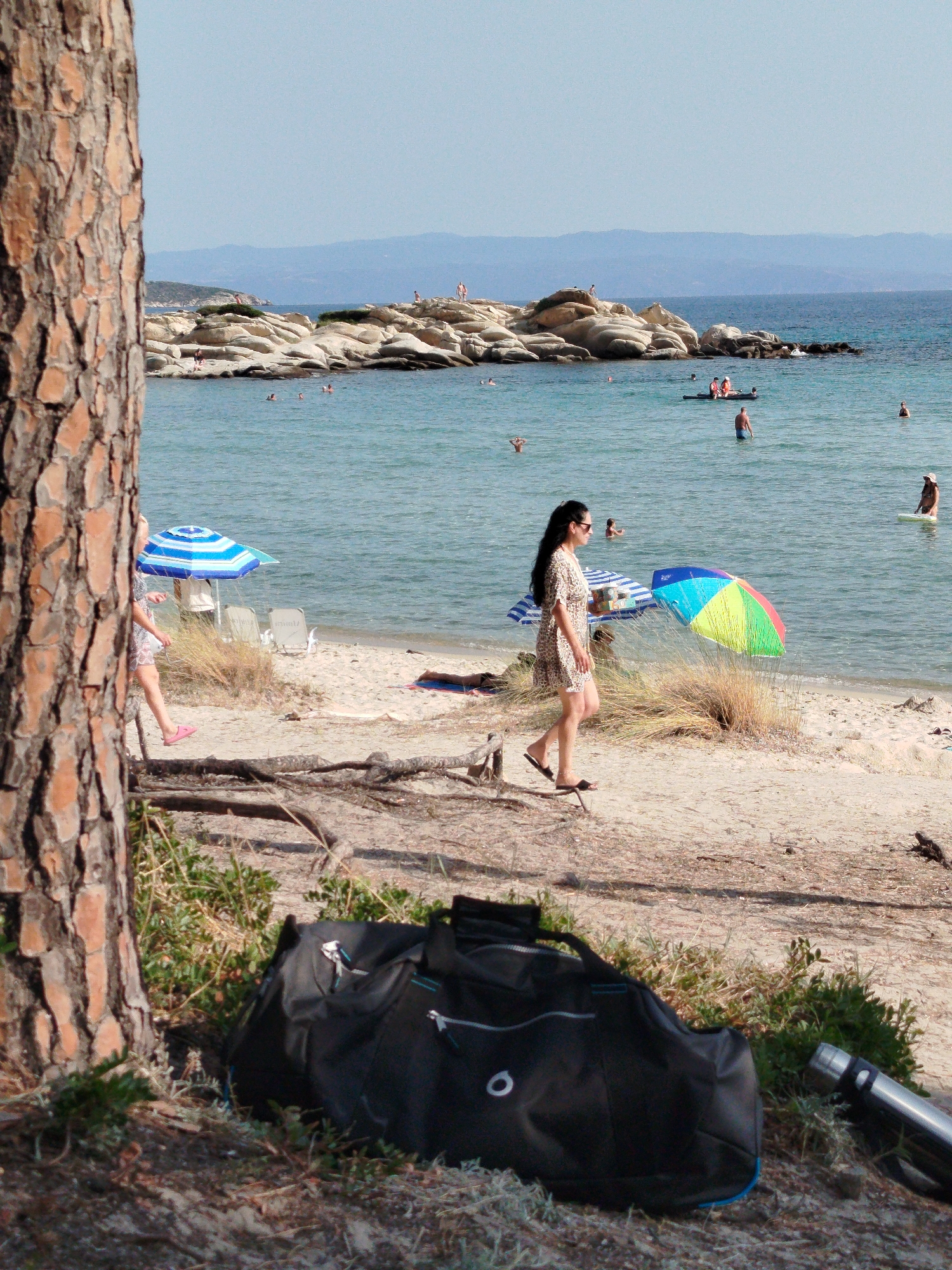 This is a medium shot of a beach scene in Sithonia, Greece, with a large tree trunk in the foreground on the left. A woman in a patterned dress and sandals walks on the sand in the mid-ground. She is facing left. Behind her, a colorful umbrella is set up, along with a striped umbrella and two lounge chairs. The sea is calm and blue, with several people swimming or wading. In the distance, rocky islands and hills are visible. In the foreground, a black duffel bag and a metal water bottle are on the ground.