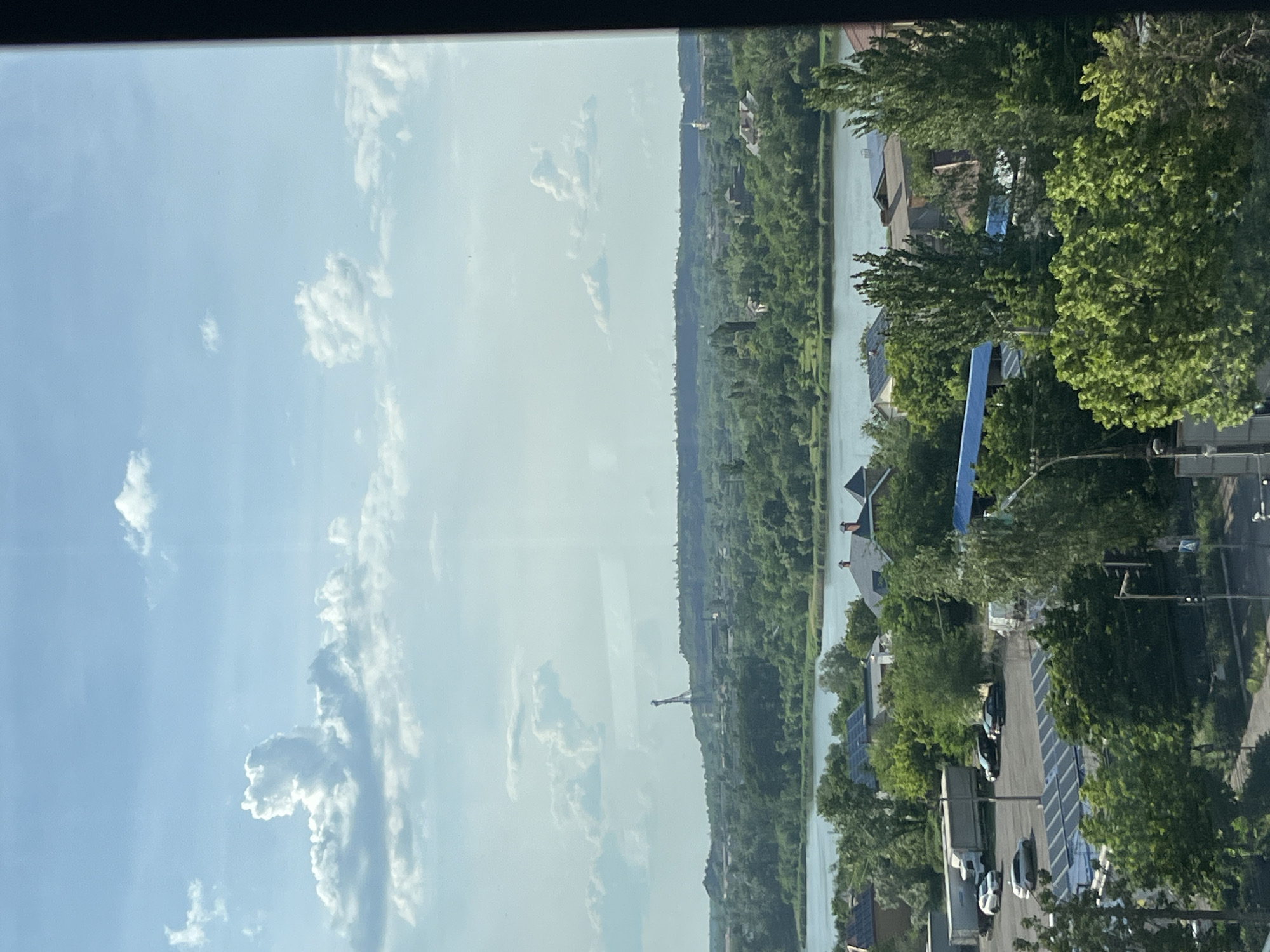 The image shows a view of a landscape from a high vantage point, likely a window. The sky is partially cloudy. Below, there's a stretch of land heavily covered with trees, bordering a body of water. On the right side of the image, there is a road with parked cars and buildings with solar panels on the roof. A utility pole is also visible. There is a crane in the background. The location appears to be near the city of Kryvyi Rih, Ukraine, given the vegetation and buildings.