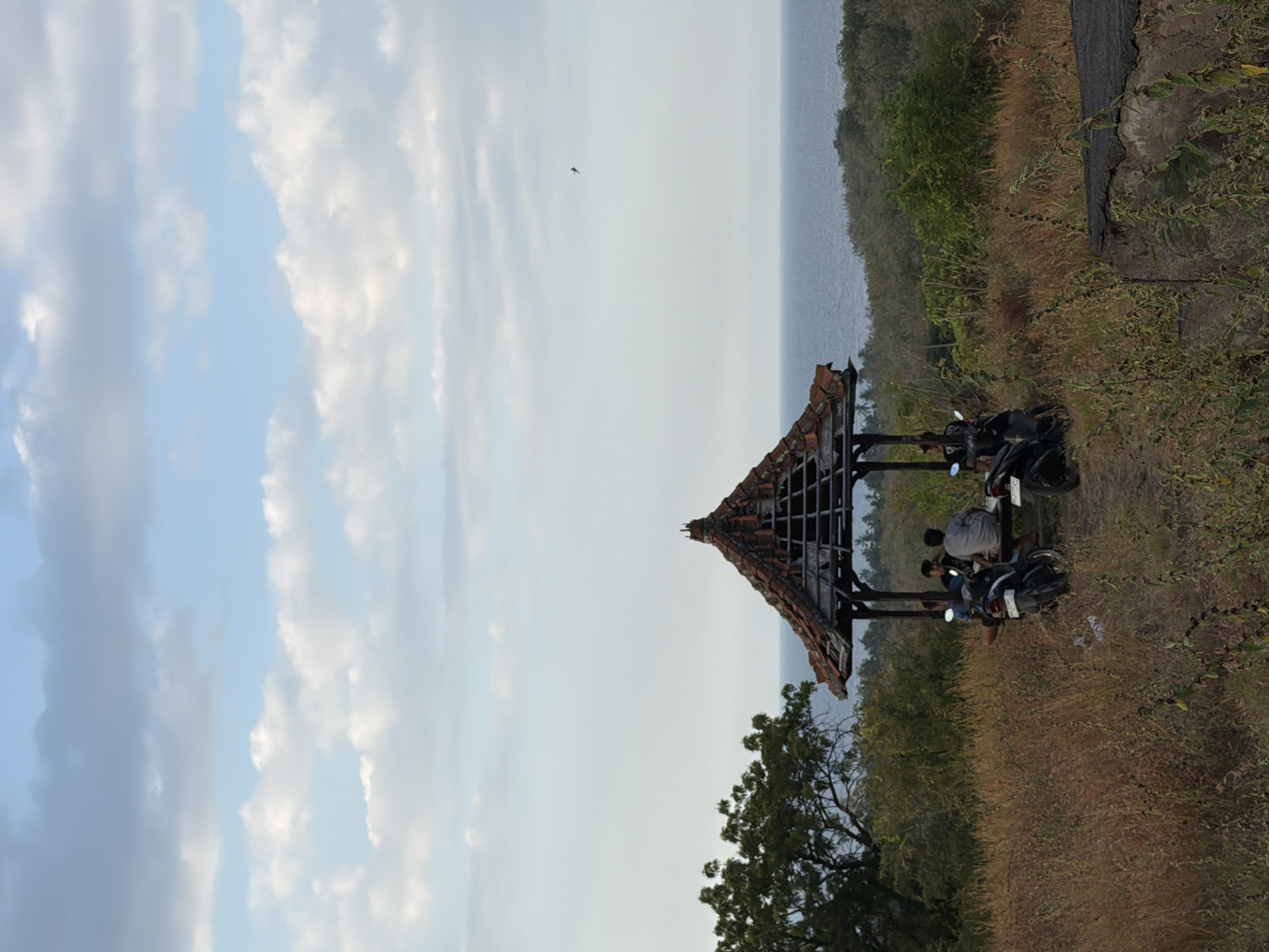 This is a vertical, eye-level, medium shot of a gazebo with a dilapidated roof in Kubutambahan, Indonesia. The gazebo is on a grassy hill overlooking the ocean. Several people are seated inside the gazebo, and motorcycles are parked outside. The sky is partly cloudy, and the ocean is visible in the background. The foreground is filled with dry grass and some green bushes. The overall atmosphere is serene and slightly melancholic due to the weathered appearance of the gazebo.