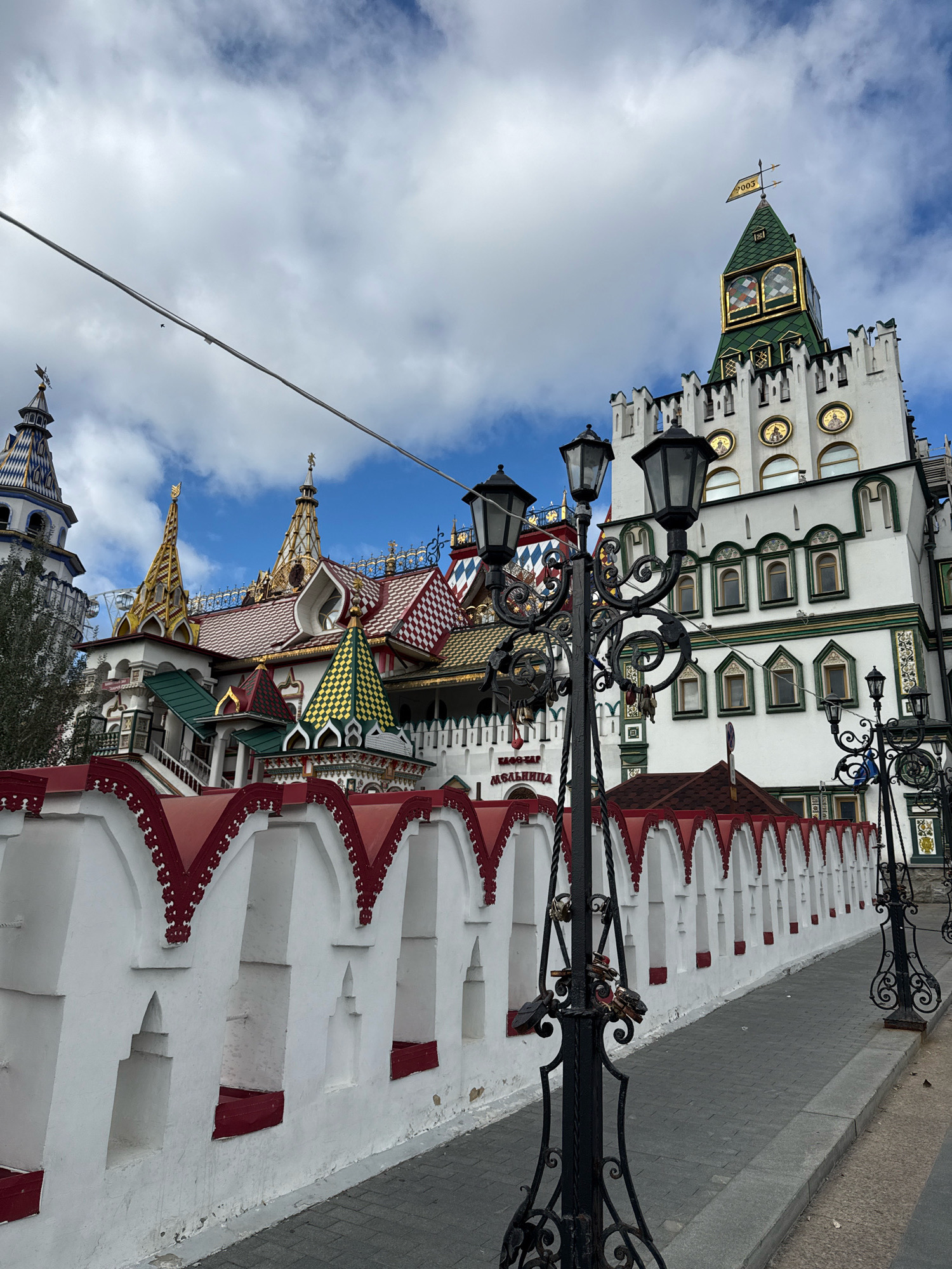 This is a photograph of the Izmaylovo Kremlin in Moscow, Russia, taken on a cloudy day. The complex features brightly colored, fairytale-like architecture with multiple towers and ornate roofs in shades of green, gold, red, and white. A white stucco wall with red decorative trim and arched openings runs along the foreground, creating a pathway. Several black, wrought-iron lampposts with decorative scrollwork and padlocks adorn the path. The Izmaylovo Kremlin is a unique cultural and entertainment complex designed to resemble a medieval Russian fortress.