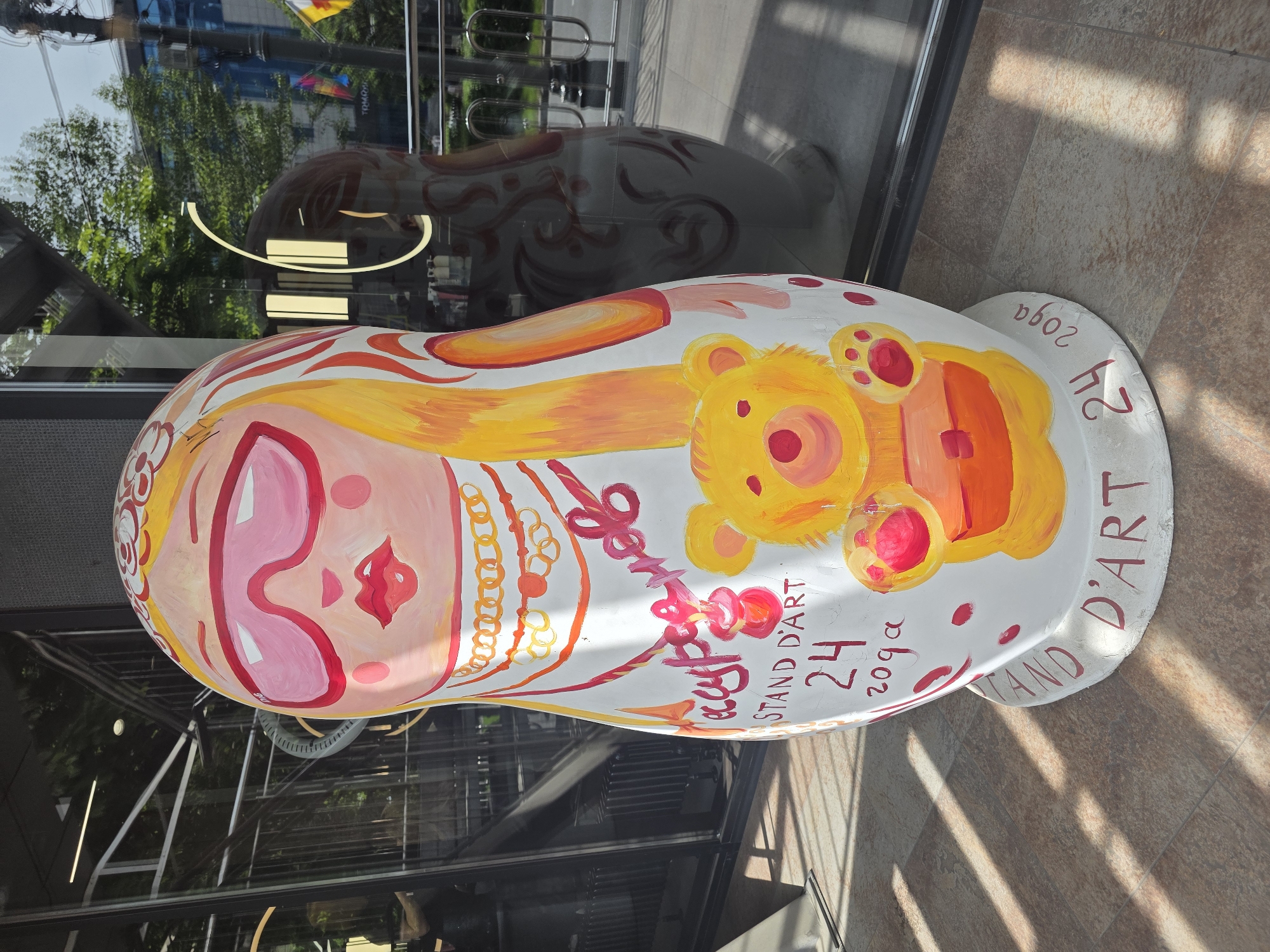 The image shows a large, painted Matryoshka doll sculpture. The doll is white with a painted face featuring pink sunglasses and red lips. It also has yellow hair, a gold necklace, and a cartoon bear image on its side. Script reading "Stand D'Art 24 2029" is visible, although the date appears to be intentionally misspelled. The doll is placed outdoors in front of a glass storefront, with reflections of trees and buildings visible in the glass. The ground beneath the doll is tiled. The sculpture’s style suggests it is part of a public art installation. The presence of the “Stand D’Art” branding could indicate the exhibit’s name. It is located in the city of Krasnodar, Russia.