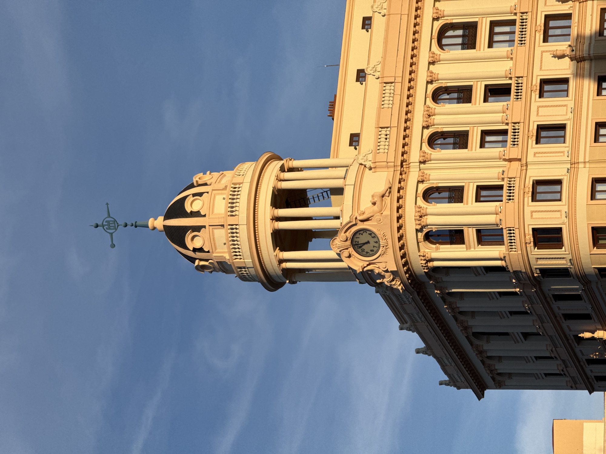 This is a photograph of the Metropolis Building, located at the corner of Calle de Alcalá and Gran Vía in Madrid, Spain. The image focuses on the building's dome, clock, and upper façade. The dome is decorated with architectural details and topped with a statue. The clock is visible on the side of the dome. The building's façade is ornate, featuring rows of windows and decorative elements. The sky is blue with some light clouds.