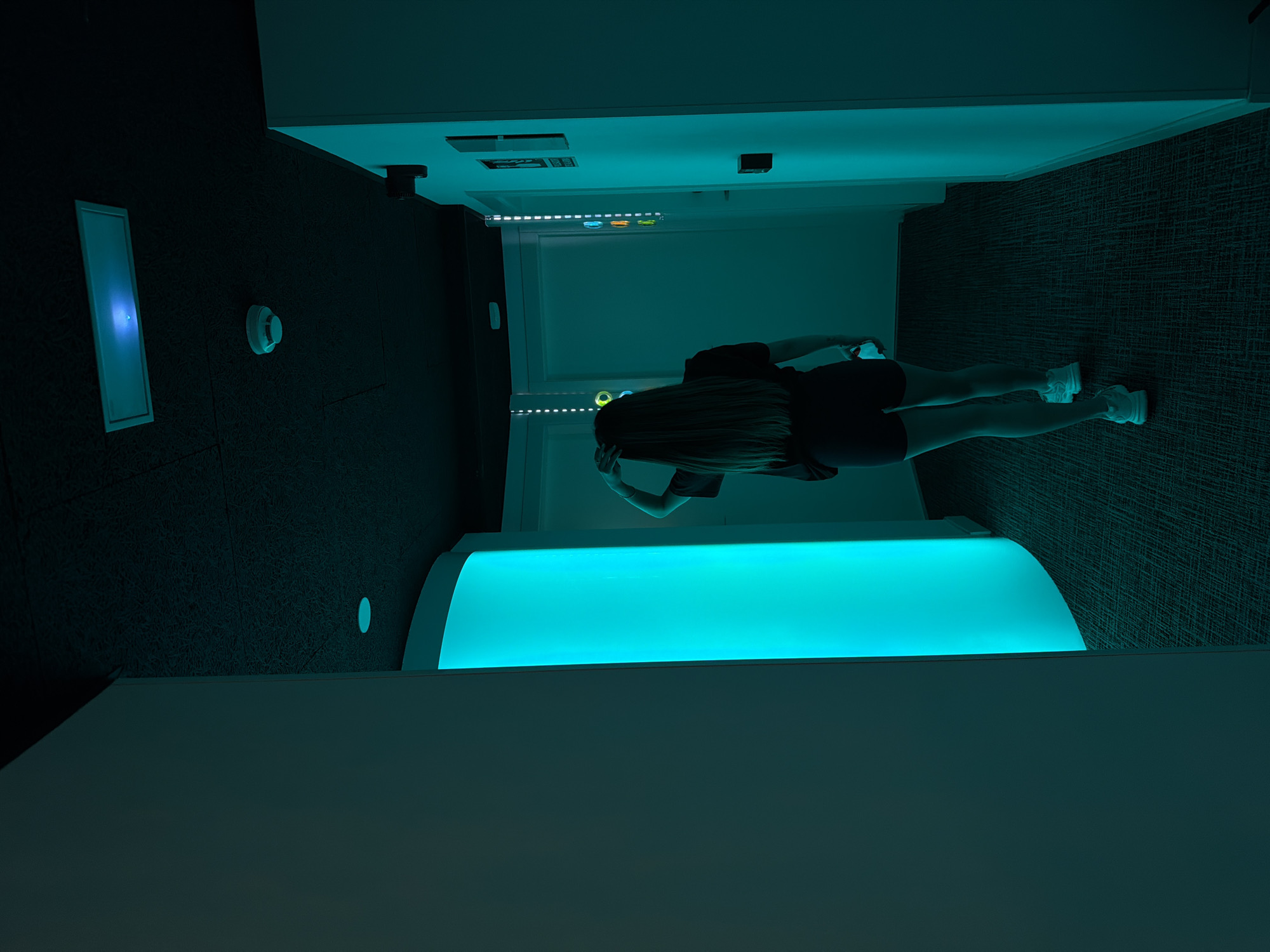 The image shows a rear view of a woman with long, dark hair walking down a dimly lit hallway. She is wearing a dark t-shirt and dark shorts, along with white sneakers. The hallway has a dark carpeted floor and white walls. To the left of the woman, there is a curved structure illuminated with bright turquoise light. The ceiling is dark and appears to have acoustic tiling. Several light fixtures and a smoke detector are visible on the ceiling. On the wall to the right, there are doors and some decorative elements, including vertical strips of lights with colored circles and a sign with a pictogram. The overall ambiance is modern and somewhat futuristic, with the lighting playing a significant role in the scene. The location is not specified in the image, and there is no indication that it is in Madrid, Spain.