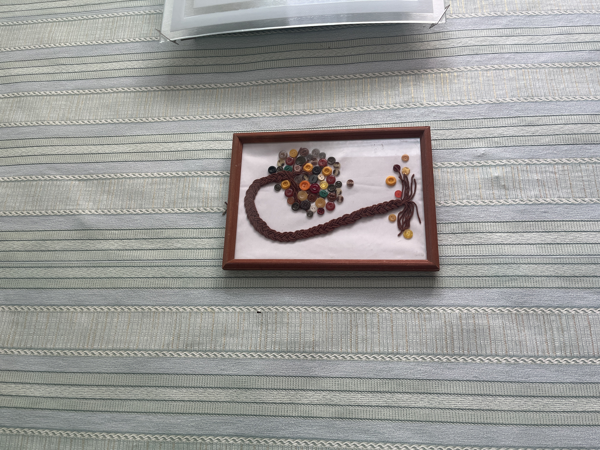 The image shows a framed artwork on a wall with vertical striped wallpaper. The artwork is a braided brown element resembling a pigtail, with a cluster of colorful buttons at the end. There are also a few loose buttons scattered around the artwork and on the wall. The wall features a pale blue and white striped pattern. To the right of the artwork, a sliver of a white object with a metallic frame is visible, possibly another decorative item or part of the room's architecture. The scene is indoors, suggesting a domestic setting. The city of Kovriga, Russia, is not depicted in the image.