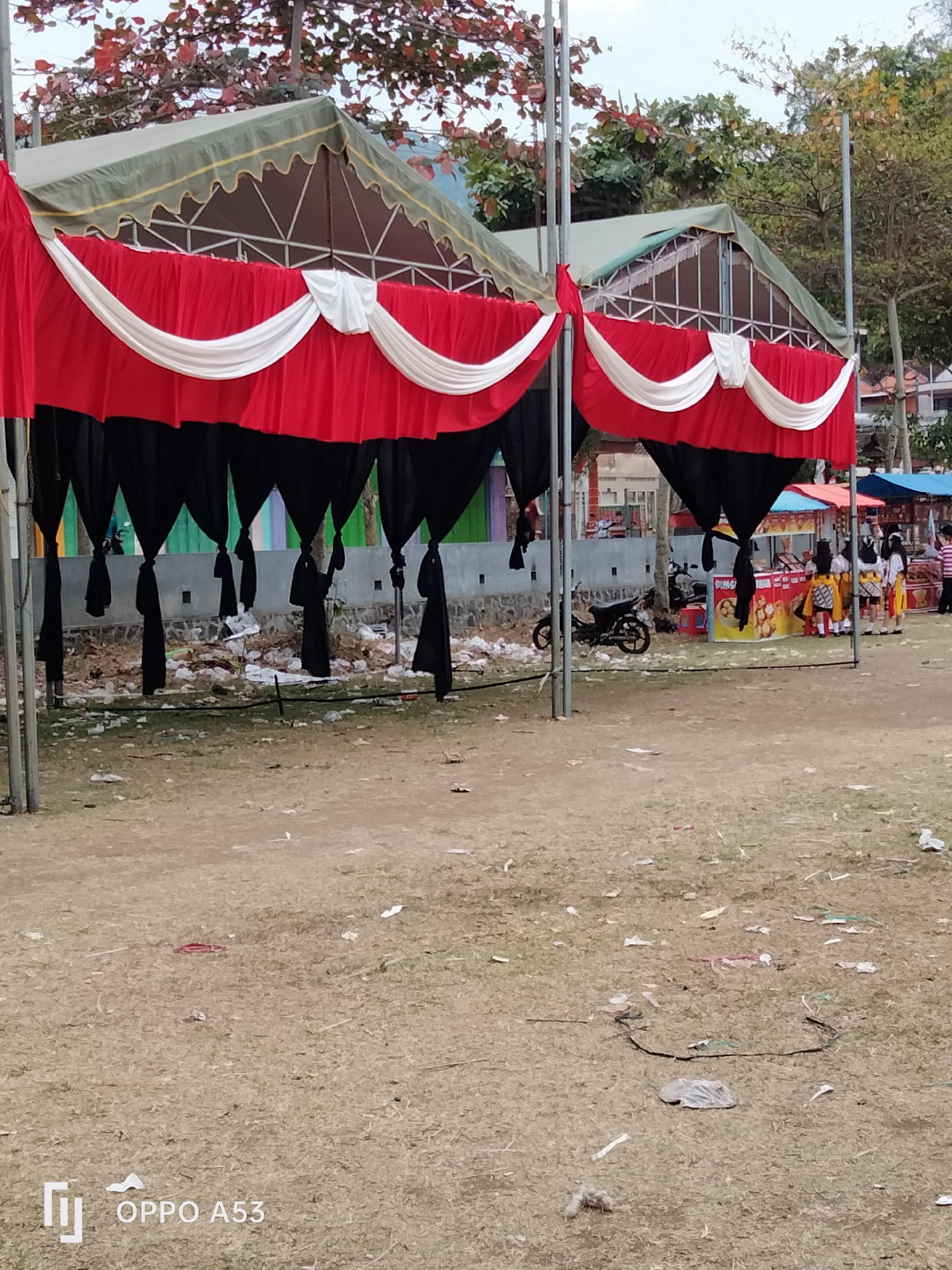 The image depicts an outdoor scene in Bendoagung, Indonesia, featuring a large tent structure with red, white, and black fabric draping. The ground is covered in dry grass and scattered litter, suggesting a public space that may have recently hosted an event or is a casual gathering area.
In the background, there are several market stalls with vendors and customers. A motorcycle is parked near one of the stalls. People, some dressed in traditional attire, are visible interacting at the stalls, with one group appearing to be young women in uniform. The overall atmosphere suggests a lively market or community event. The presence of multiple tents and stalls indicates a commercial or festive environment. The condition of the ground with litter points to a busy area, possibly with limited cleanup.