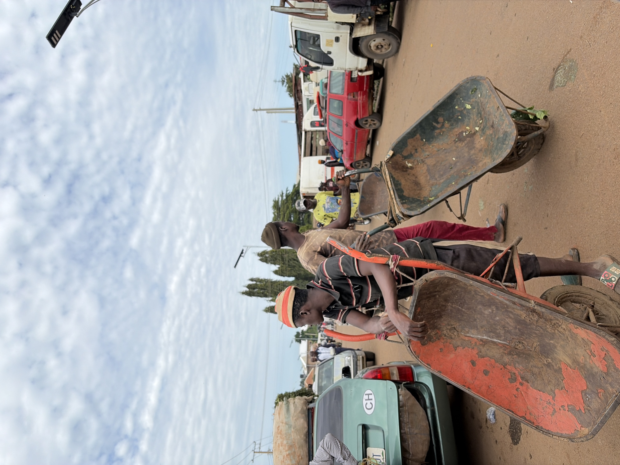In Gura Top, Nigeria, a street scene unfolds with a sky filled with white clouds. Two men are prominent in the foreground, each associated with a wheelbarrow. One man, wearing a striped shirt and an orange and white hat, is actively pushing a red wheelbarrow. The other man, in a brown shirt and a dark hat, is near a green car and appears to be interacting with the wheelbarrow. In the background, various vehicles are parked and in motion, including a white truck, a red car, and other vehicles that are partially obscured. The ground is paved with reddish-brown material. The overall impression is of a busy street, possibly a market or a transportation hub, with people engaged in work or commerce.