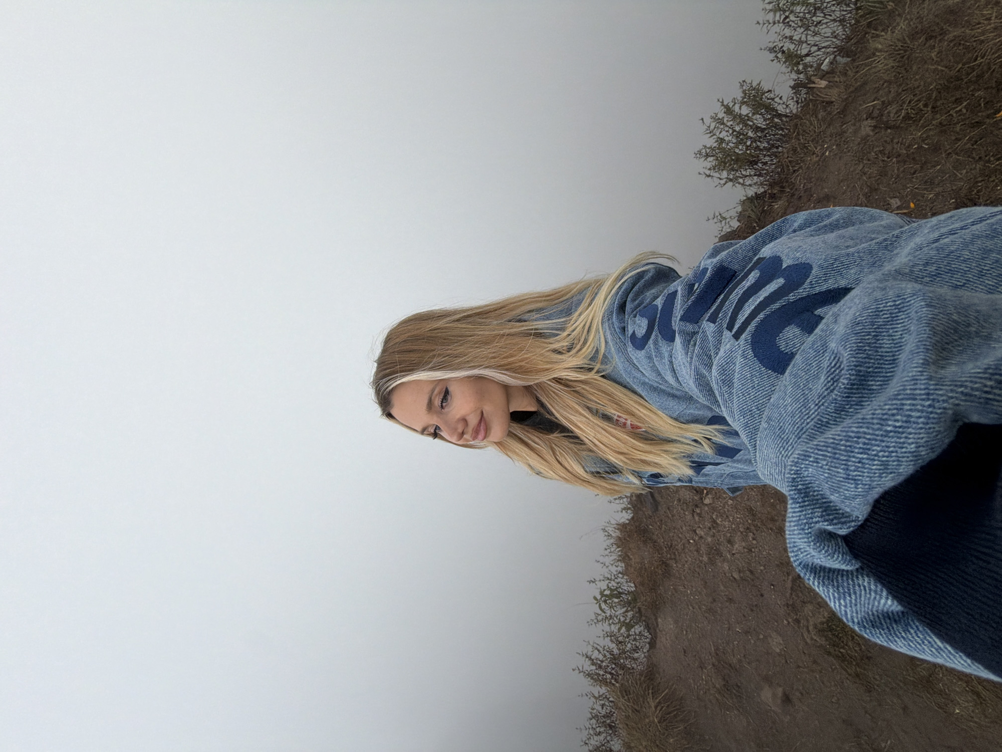 A woman with blonde hair is taking a selfie on a dirt path with sparse vegetation, possibly in Kintamani, Indonesia. She is wearing a denim jacket with blue lettering. The background is obscured by thick fog, indicating a cloudy or misty atmosphere. The ground appears to be dry and rocky, with some dry grass and small bushes growing on it. The woman is looking off to the side with a slight smile, her hair is long and flows down her shoulders.