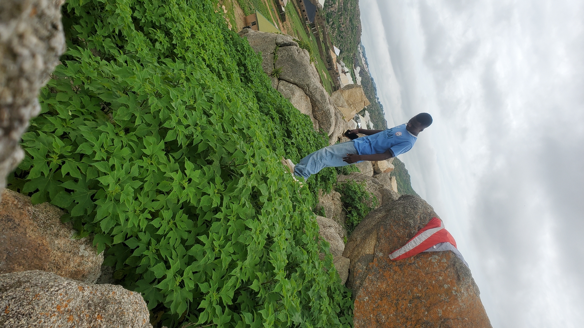 A young Black man wearing a blue t-shirt and light blue jeans stands on a rocky, vegetation-covered hillside in Jos, Nigeria. He is looking down, appearing to be holding a phone in his right hand. To his right, a large boulder with orange and gray lichen is partially covered by a red and white jacket. The hillside is dense with green foliage. In the background, a settlement with various buildings and roofs is visible on other hills. The sky is overcast with gray clouds.