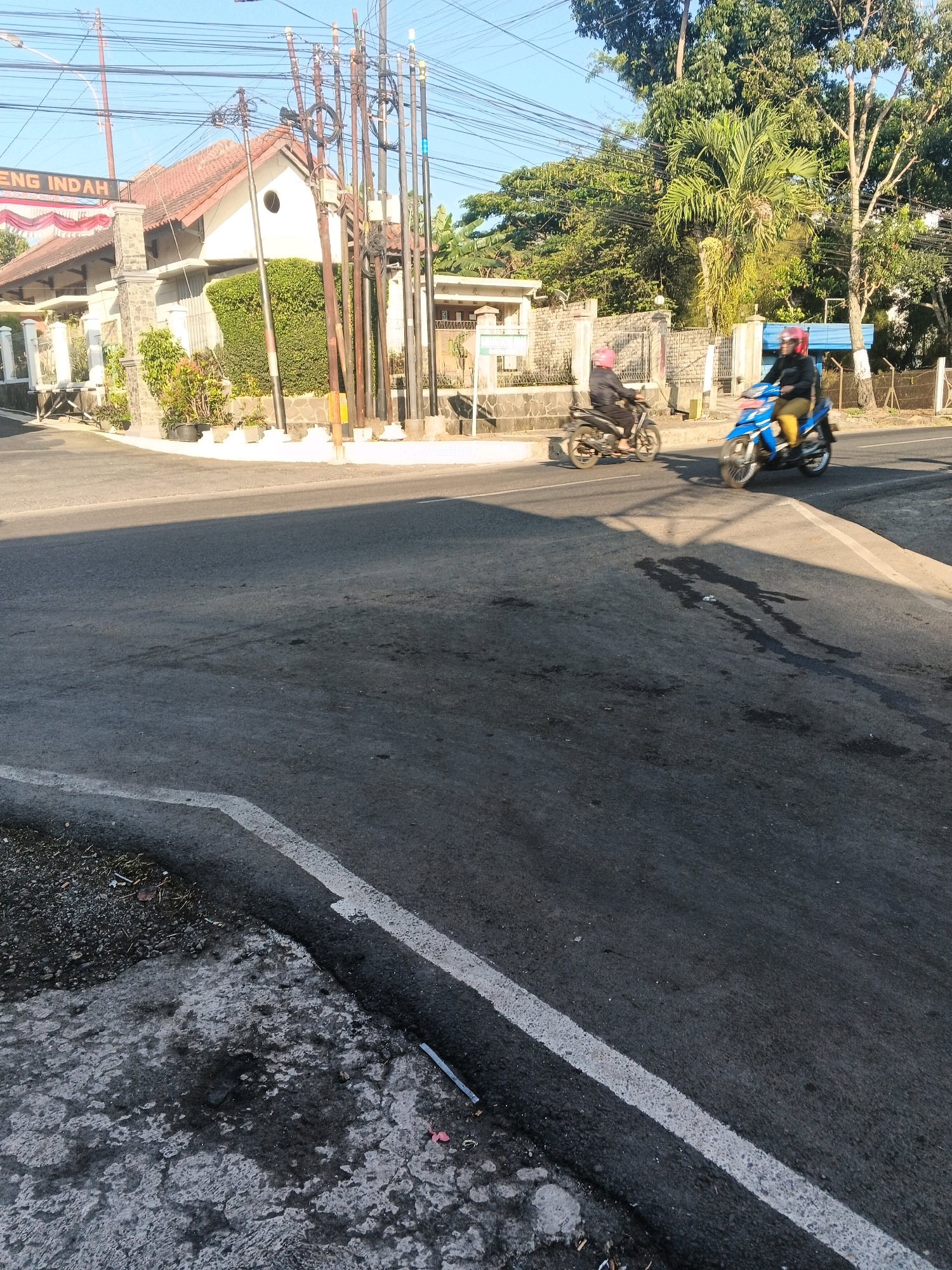 The image shows a street intersection in Semarang, Indonesia, on a clear, sunny day. Two motorcyclists are visible, one on a blue motorcycle and the other on a darker motorcycle, both wearing helmets. The blue motorcycle is in the foreground, moving away from the viewer, while the darker motorcycle is further back and to the left. The street is paved, with a white line marking the edge of the lane. On the left side of the street, there is a building with a red-tiled roof and a sign that reads "ENG INDAH." Behind the building, there are trees and utility poles with many wires. The right side of the street has more trees and some fencing. The lighting suggests it is either early morning or late afternoon, with shadows cast across the road. There are some oil stains and debris on the road surface.