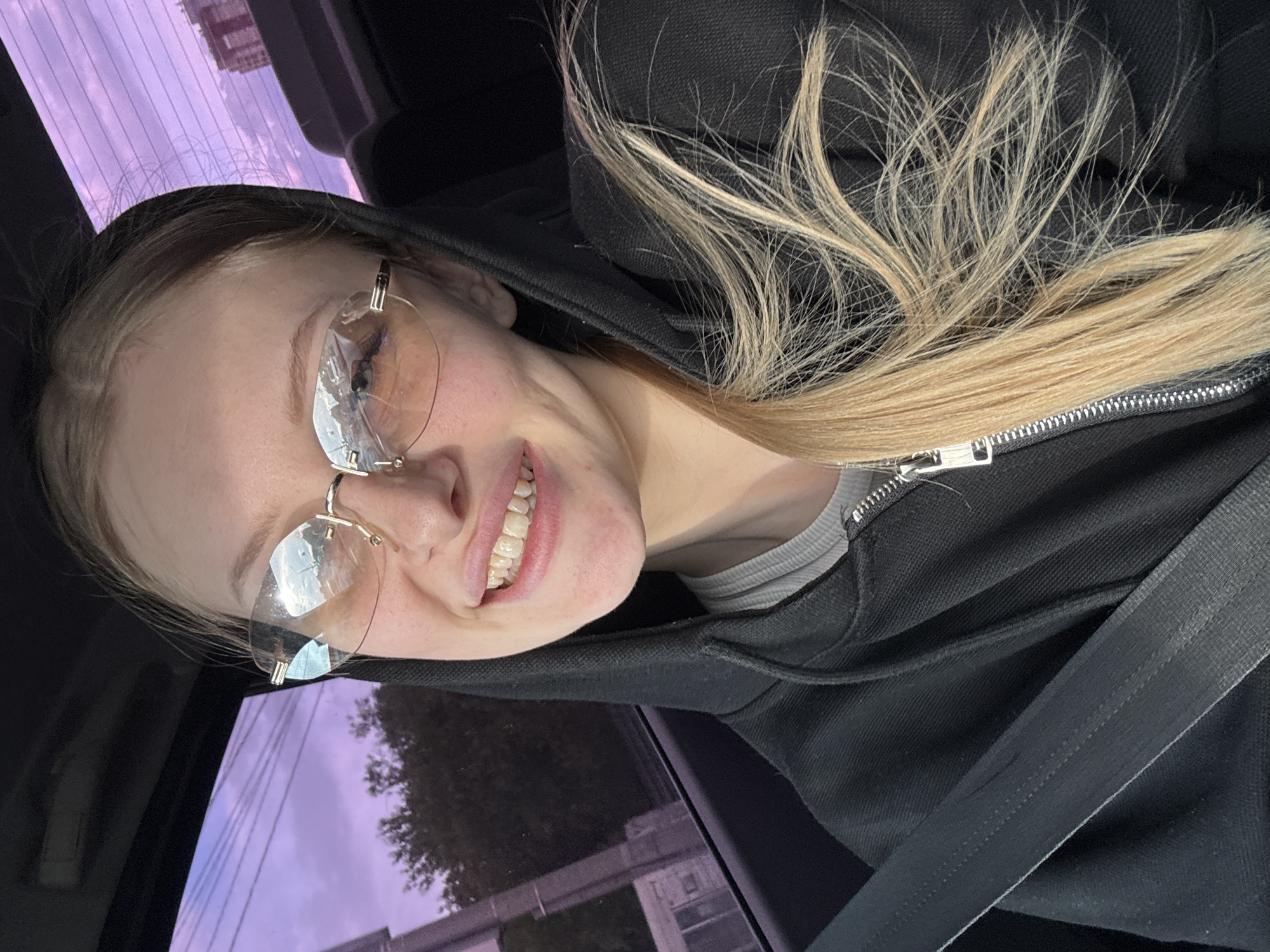 A woman is in the driver's seat of a vehicle. She is wearing glasses and a black hooded sweatshirt. The car is in motion, as evidenced by the blurred cityscape visible through the car window. While the exact location within Perm, Russia, cannot be definitively determined from the image, the blurred background suggests an urban setting with buildings and possibly trees. The sky appears to be at dusk or dawn, with hues of pink and purple. The woman is smiling and appears to be looking at the camera. A seatbelt is visible across her chest.