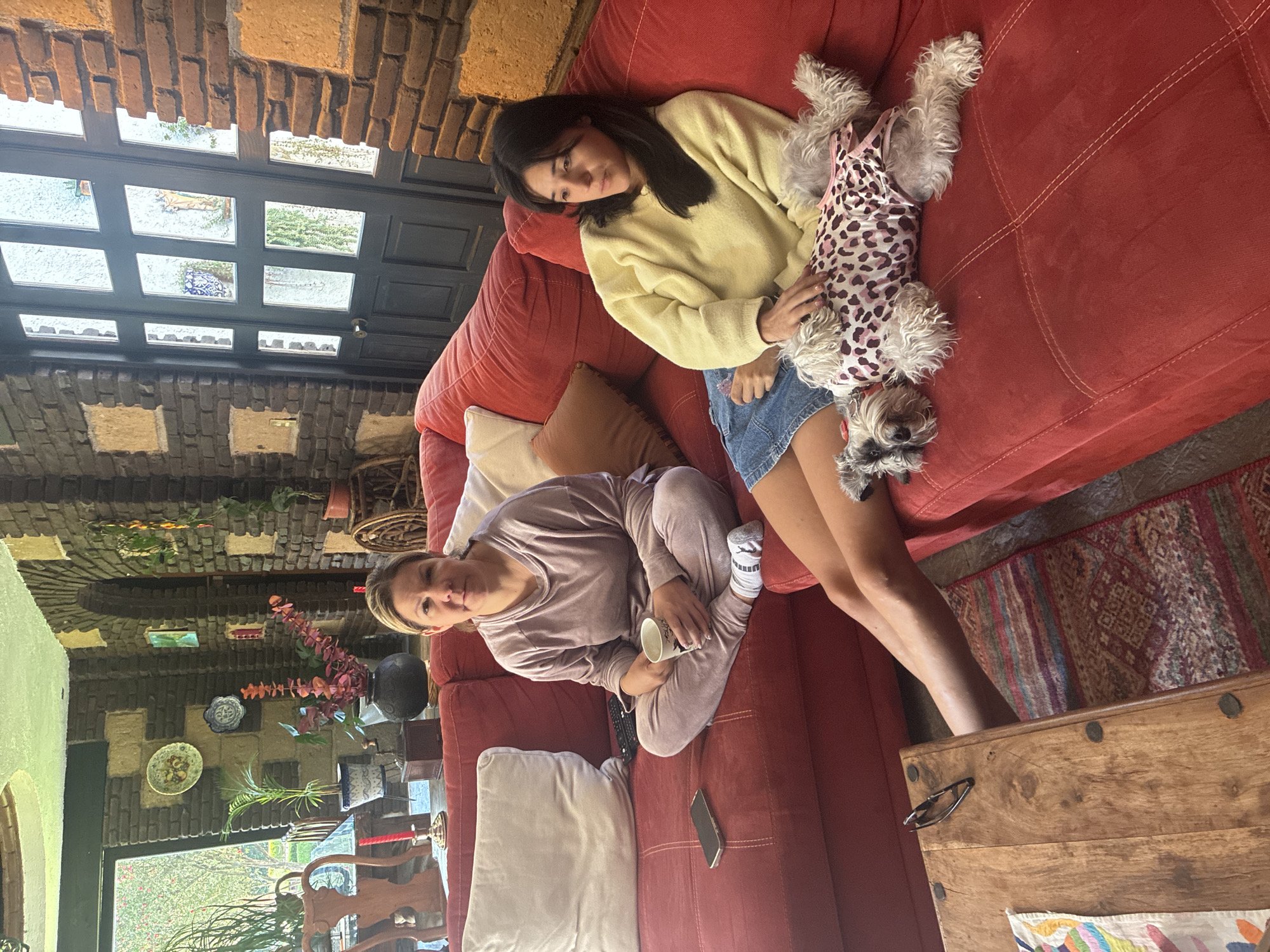 The image shows two women and a small dog on a red couch in a room with unique architecture. One woman is wearing a light yellow sweater and denim shorts, reclining and holding a small dog in a leopard print outfit. The other woman is seated and wearing a light purple pajama set, holding a mug. A smartphone sits on the couch near the seated woman. The room has a brick wall and ceiling with glass inserts that offer a view of the outdoors. In the background, a window reveals a garden or patio. The location is Cañada de Madero, Mexico. A decorative rug is visible near a wooden chest.