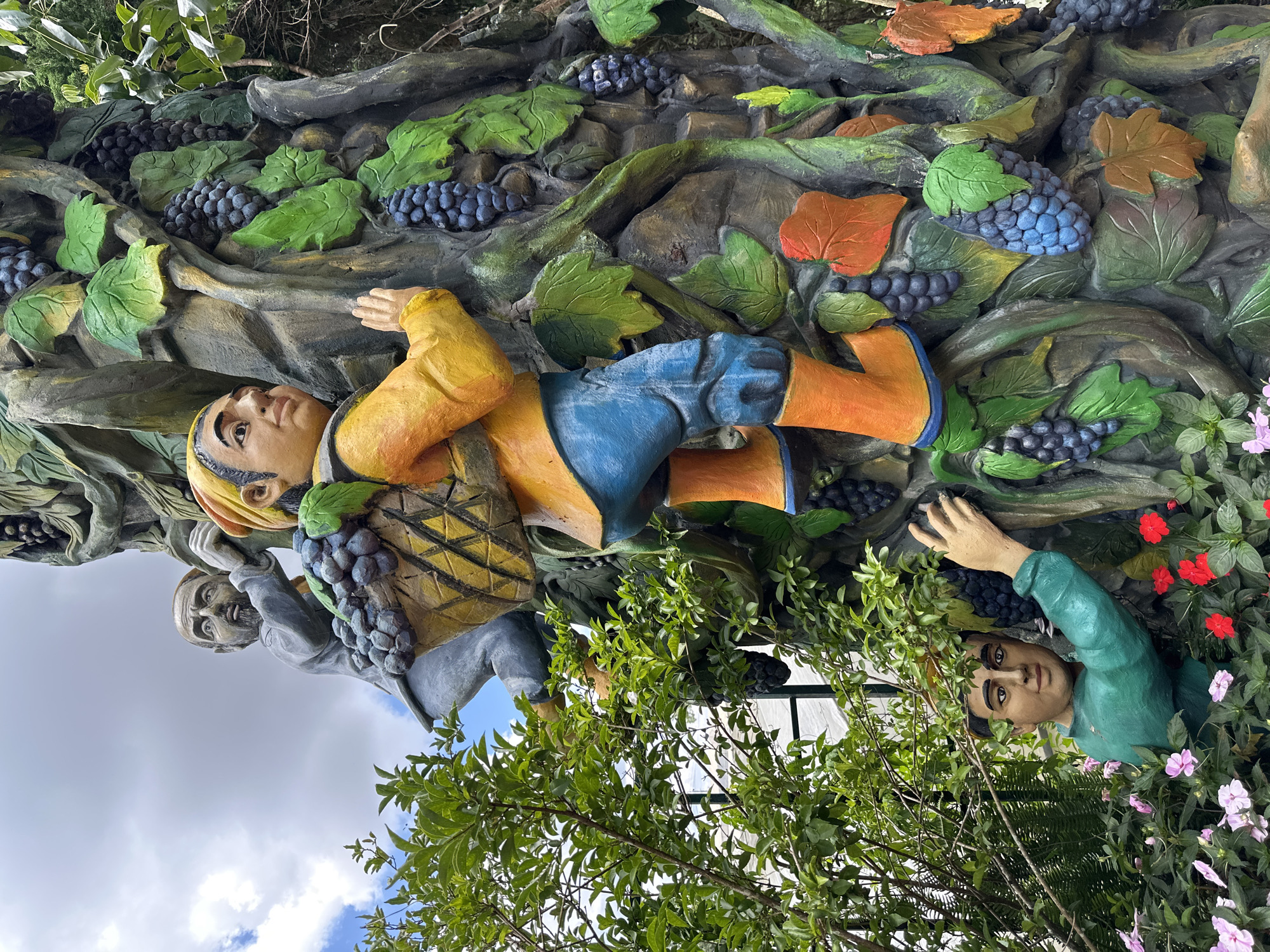This image depicts a colorful, textured sculpture of men harvesting grapes from a large, stylized vine. The sculpture is located in Da Lat, Vietnam, specifically at the "Valley of Love" attraction. In the foreground, a man in a green shirt is partially obscured by lush foliage and flowers, reaching towards a bunch of grapes. Above him, a second man, dressed in yellow and blue with orange boots, is climbing the vine, carrying a woven basket filled with grapes on his back. His posture suggests exertion and focus on his task. Higher up on the vine, a third man, with a grey shirt and a beard, is also climbing, his hand gripping the vine for support. The sculpture is detailed with faux grapes, leaves, and vines, creating a vibrant and engaging scene. The background shows a bright, cloudy sky and more greenery, indicating an outdoor setting in a park or garden.