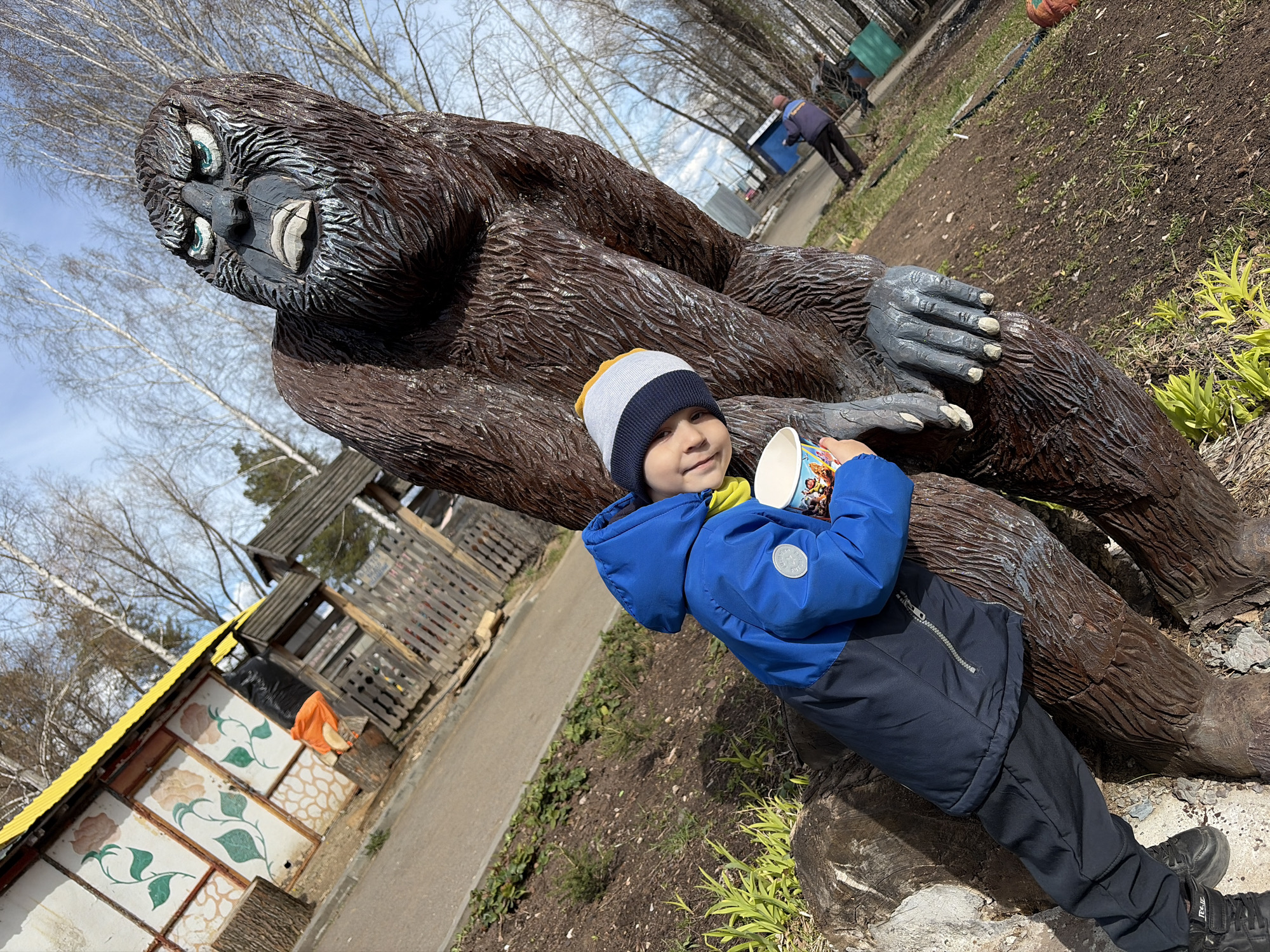 A child in a blue jacket and hat is standing next to a large, brown, wooden statue of a yeti or bigfoot in a park. The child is holding a cup with characters printed on it. The statue is laying down with its arm outstretched. In the background, there is a building with floral paintings, trees, and other people. The location is likely in Chaikovsky, Russia.