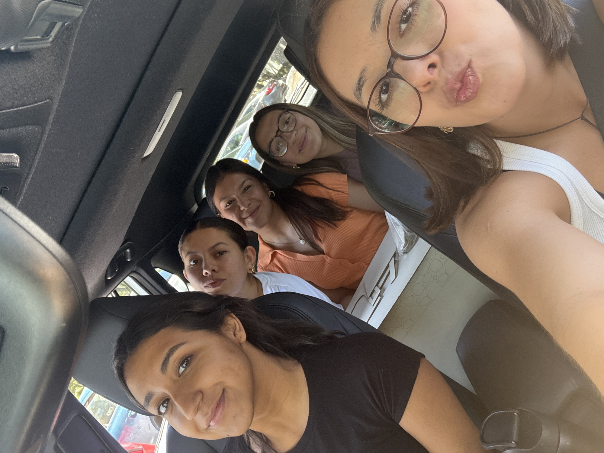 Five young women are crammed inside a vehicle, likely a car, as they pose for a selfie. The photo is taken from an elevated angle, as if by someone in the front passenger seat. The location appears to be San Andrés Cholula, Mexico, based on identifiable architectural styles and prevalent vegetation. The women vary in complexion and are dressed in casual clothing. One wears a white tank top and glasses, puckering her lips for the camera. Another has an orange shirt and glasses. The vehicle's interior is predominantly black.