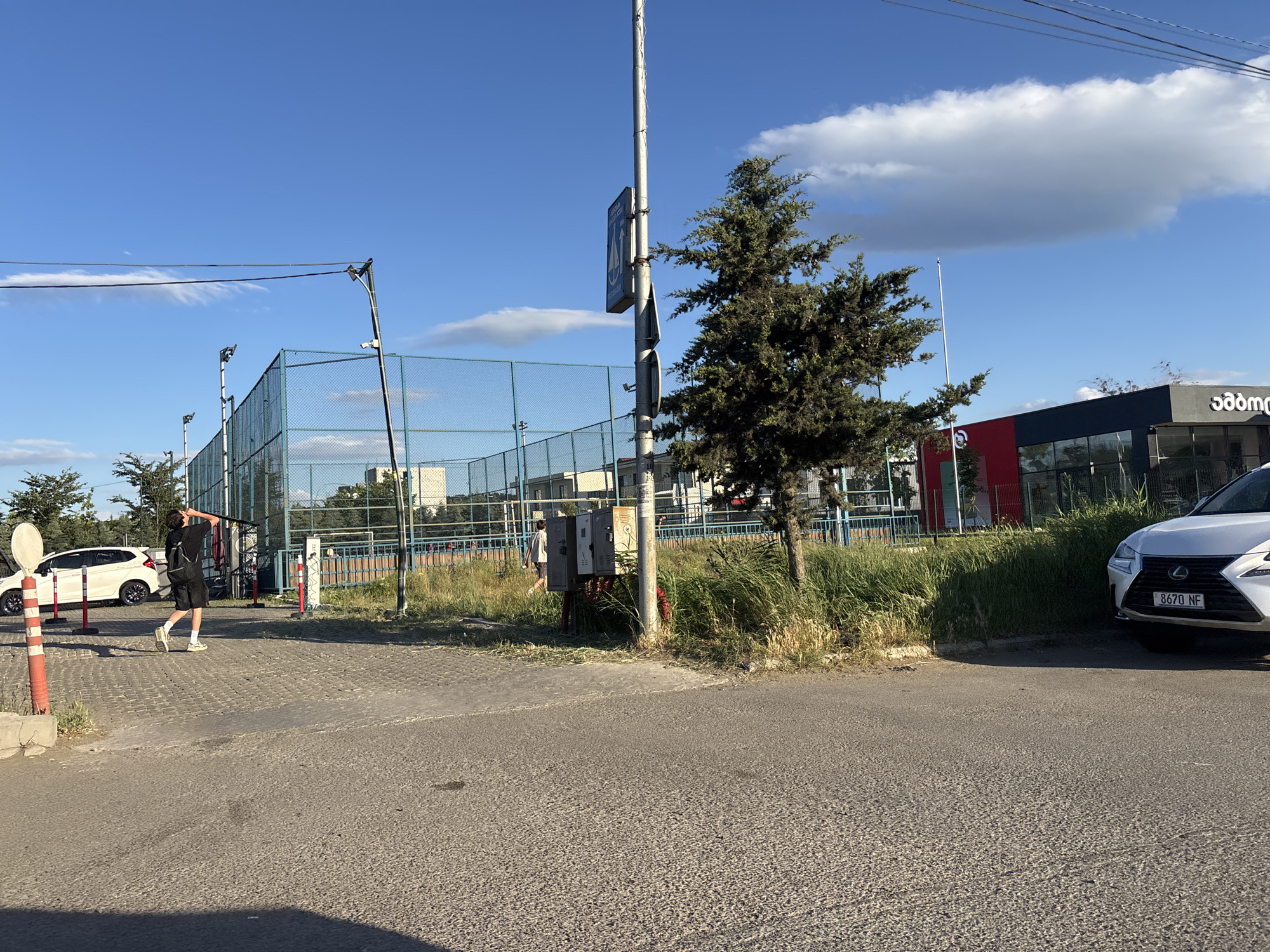 The image depicts an outdoor scene in Tbilisi, Georgia, featuring a sports court enclosed by a chain-link fence. A young person, possibly male, wearing a black t-shirt, shorts, and sneakers, is visible on the paved area outside the court, carrying a backpack and looking up. A white car is parked to the left of the frame, and a white Lexus is parked on the right. The sky is blue with scattered clouds. The foreground shows a paved road, and the background includes a building with signage in Georgian script, a tree, and other vegetation. The overall atmosphere suggests a sunny day.