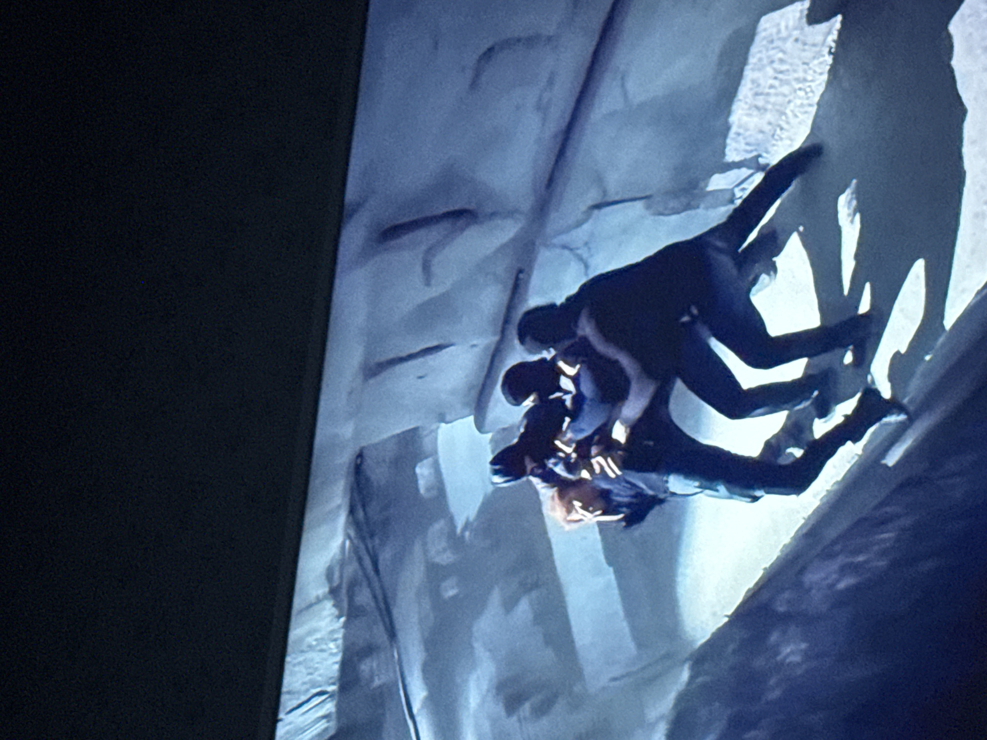 The image shows a scene from a movie, depicting three individuals engaged in a physical struggle. They appear to be positioned on a vertical or near-vertical surface, possibly a wall or cliff face, within an ambiguous environment. One individual wears lighter colored clothing with dark trim or accents, suggesting a possible distinction in attire. The actions suggest a conflict. The setting is not clearly identifiable. The location in the city of Mytishchi, Russia is unknown.
