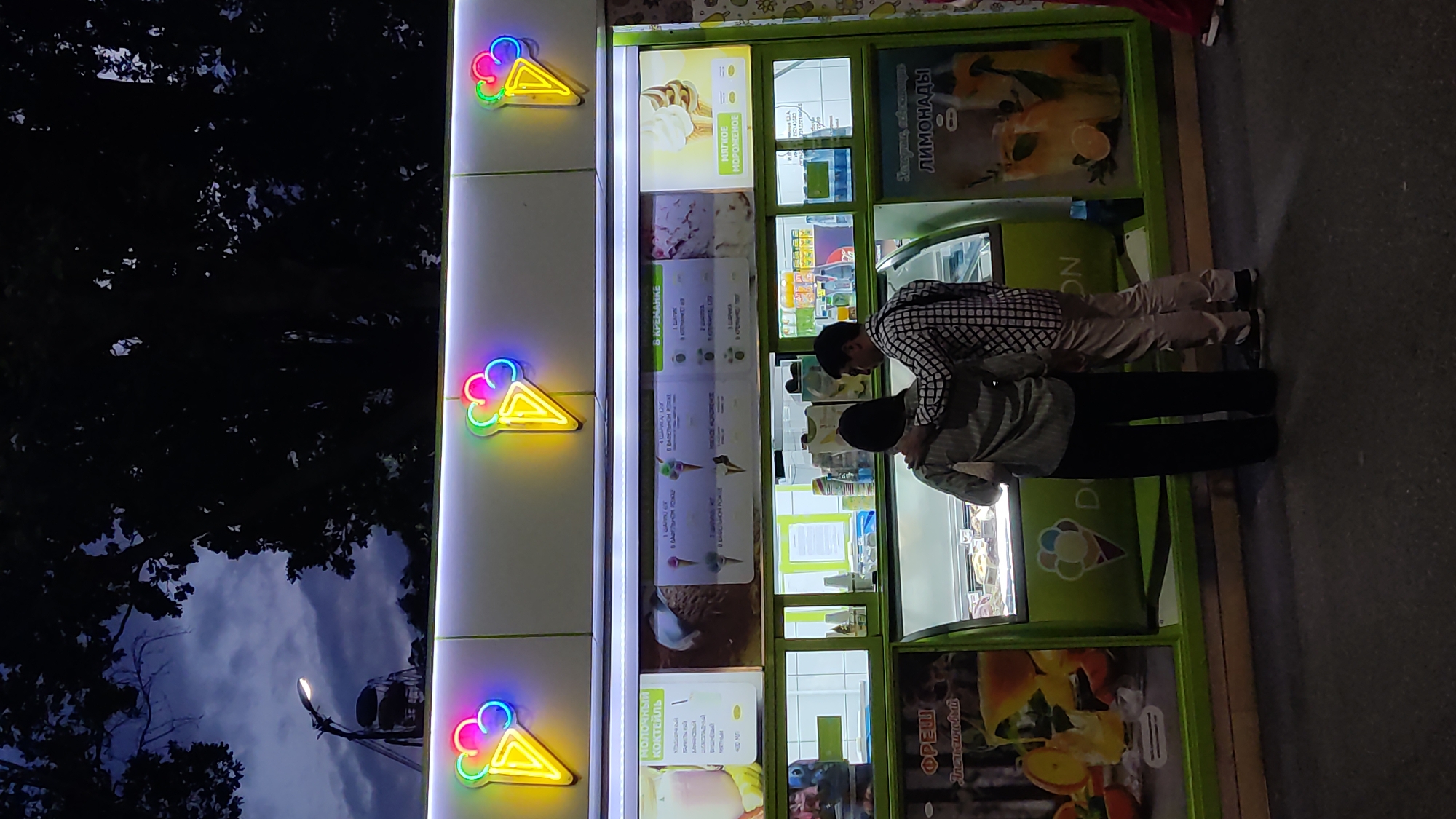 In Krasnodar, Russia, at dusk, an ice cream stand is illuminated by neon signs shaped like ice cream cones. A couple, a man and a woman, stand in front of the stand, with the man's arm around the woman's shoulders. The stand displays various ice cream flavors and lemonade. The background shows trees and a cloudy sky.