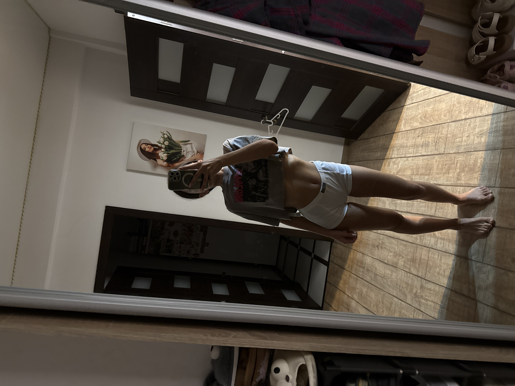 A young woman is taking a selfie in a mirror, wearing a grey cropped t-shirt and white boy shorts. She is standing in a room with wooden floors and two dark wood doors in the background. To her left, a tall mirror reflects the scene, and to her right, a closet is visible with several pairs of shoes on the bottom shelf. A painting of a woman holding a bouquet of flowers hangs on the wall behind her. The lighting in the room is dim, with sunlight casting shadows on the floor.