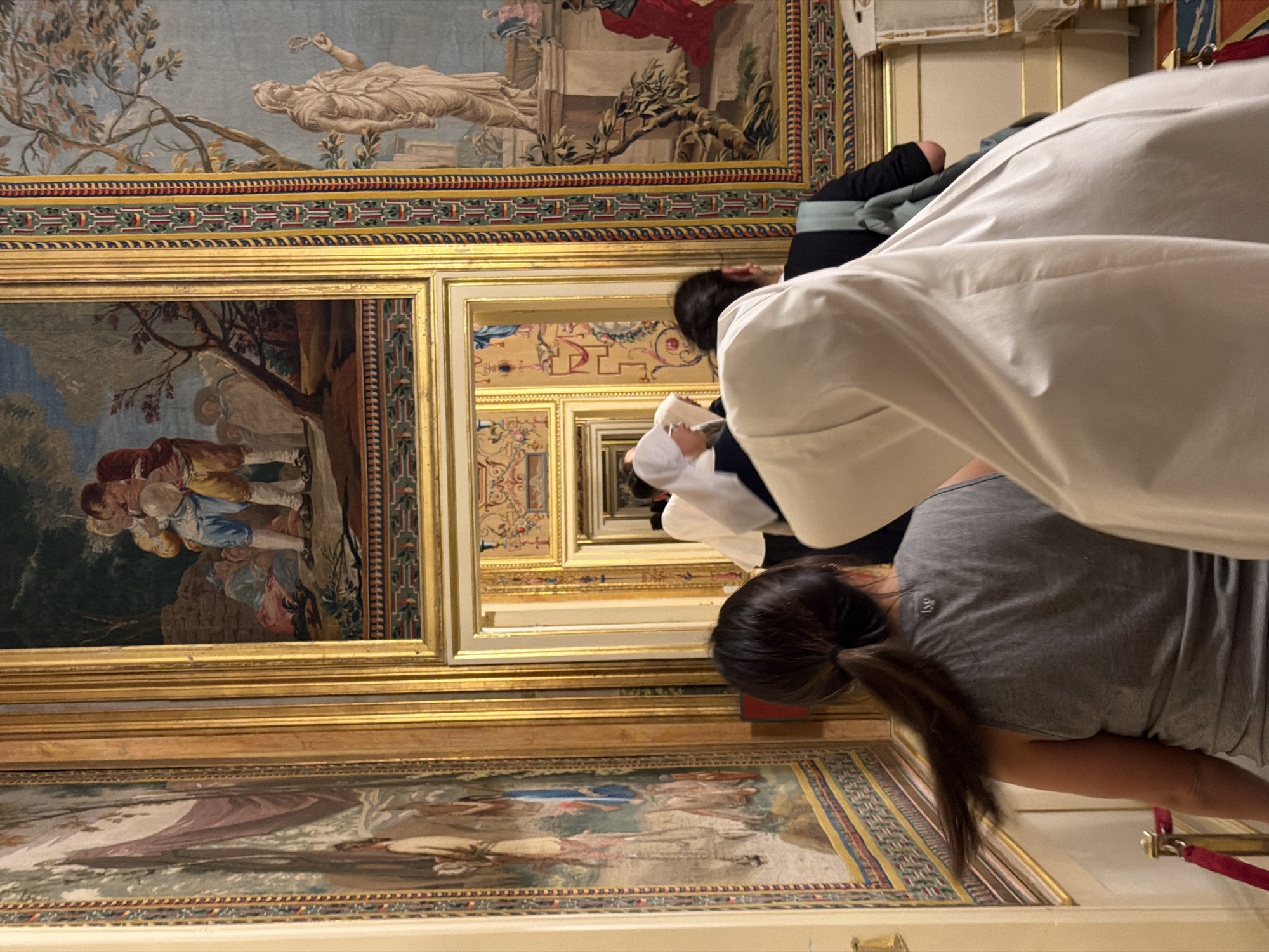 The image shows a group of people looking at tapestries in a richly decorated hall. The hall features ornate gold frames surrounding several large, colorful tapestries depicting classical scenes with figures. The tapestries are arranged in a way that creates a sense of depth, with some appearing to recede into the distance. The people in the image are observing the artwork, with one person in the foreground wearing a grey t-shirt and another in a white robe. The setting appears to be a museum or historical building, possibly the San Lorenzo de El Escorial in Spain, given the context. The lighting suggests an indoor environment, possibly illuminated by artificial lights or natural light filtering in from windows. The mood seems to be one of quiet contemplation and appreciation of the art. There is no visible text in the image.