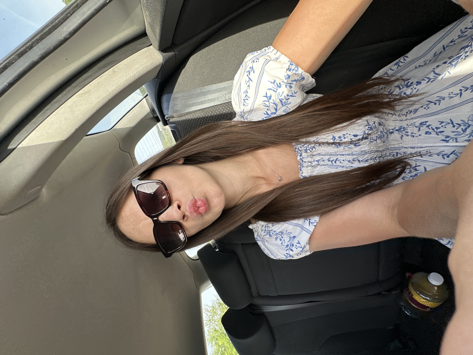 A young woman is in the passenger seat of a car. She is wearing sunglasses and a blue and white dress. The interior of the car is visible. The car is presumably in Rechtenbach, Germany. The woman is posing. A bottle of juice is on the floor of the car.