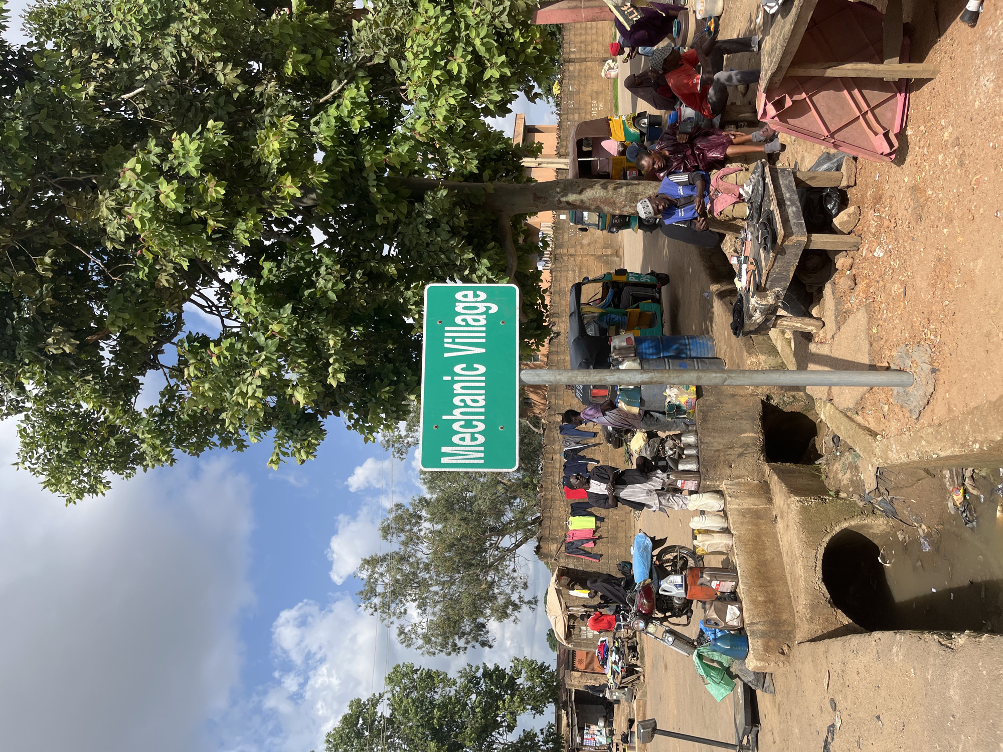 The image depicts a busy street scene in what is identified as Mechanic Village, Jos, Nigeria. A prominent green sign reading "Mechanic Village" is planted on the right side of the frame. The scene is characterized by a mix of commercial activity and daily life.
In the foreground, a concrete ditch runs diagonally across the image, filled with murky water and debris. Several containers, possibly for oil or fuel, are placed along the edge of the ditch, near a parked motorcycle and a scale.
Further back, a row of stalls and makeshift shops line the street. People are engaged in various activities. Some appear to be working, with tools and parts visible on tables. Others are seated, perhaps resting or interacting. Clothing is hung out to dry on a wall behind some of the stalls.
Several three-wheeled vehicles, commonly known as "keke napep" or auto-rickshaws, are present, indicating a form of local transportation and commerce. The background shows a brick wall and buildings, suggesting an urban or semi-urban environment. Large trees with dense foliage provide shade over parts of the scene, and the sky is partly cloudy.
The overall impression is one of a vibrant, functional marketplace where mechanical services are offered, and daily life unfolds amidst the hustle and bustle. The presence of the "Mechanic Village" sign clearly marks the location as a hub for automotive repair and related businesses.