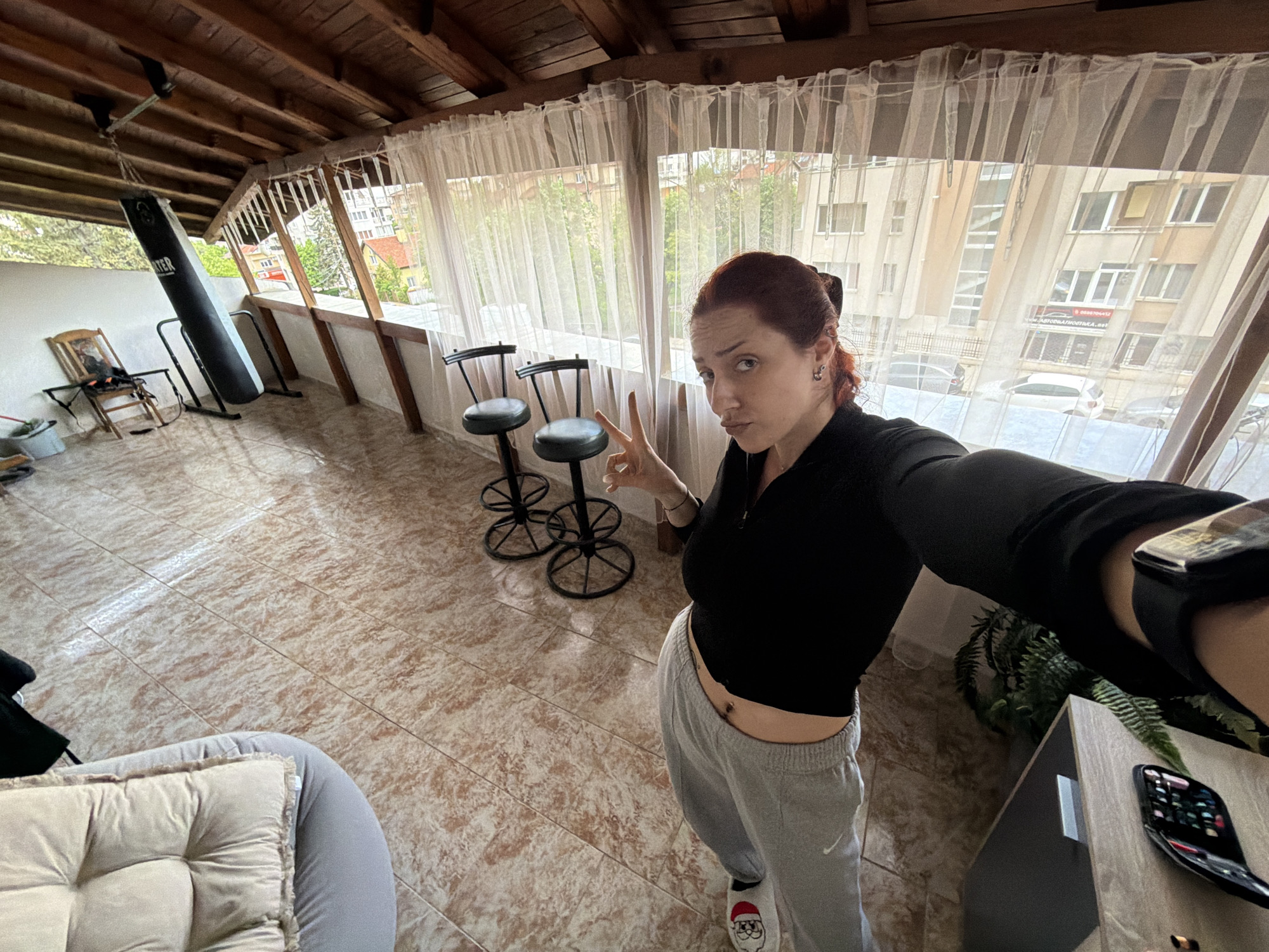 A medium shot of a woman taking a selfie in a room with a covered outdoor area. The woman has red hair pulled back, wears a black top and grey sweatpants, and makes a peace sign with her hand. She is wearing a smartwatch on her wrist. The room has a tiled floor and a sloped wooden ceiling. Behind her is a covered outdoor area with sheer white curtains. Two black bar stools are visible in the outdoor area. In the background, buildings are visible through the curtains, suggesting an urban environment. A punching bag hangs near a chair. Based on the architecture and the presence of Cyrillic script on a building sign visible through the curtain, this location is likely in Sofia, Bulgaria.