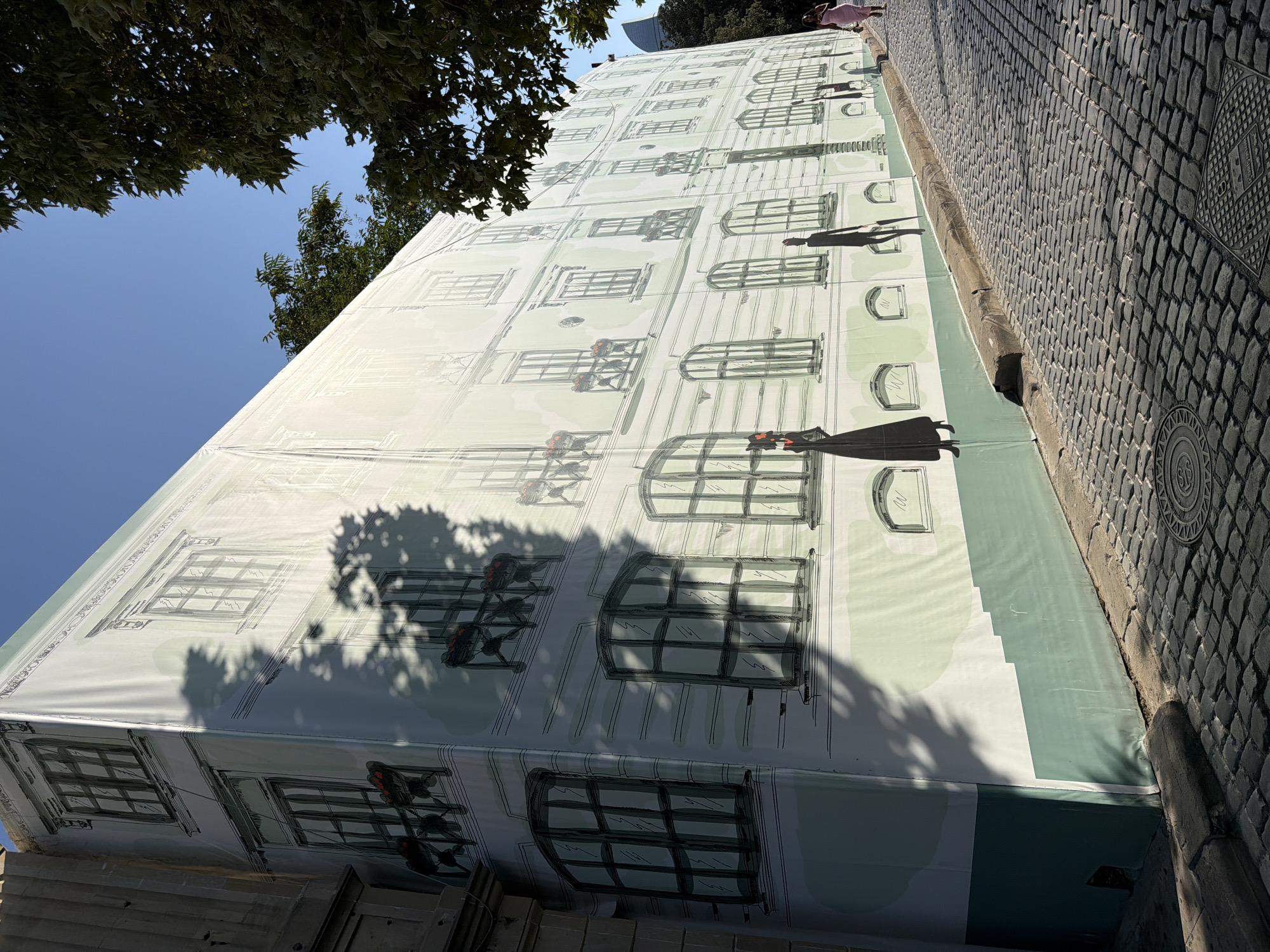 The image shows a wide, cobblestone street in Sabail Raion, Azerbaijan, with a large building covered by a white and green tarp. The tarp is decorated with a mural depicting the facade of a historic building with windows and balconies, and silhouettes of people walking. The sun is shining, casting shadows from trees onto the tarp. There are a few people visible on the street, including two women walking on the right side of the image. The architecture suggests a European influence.