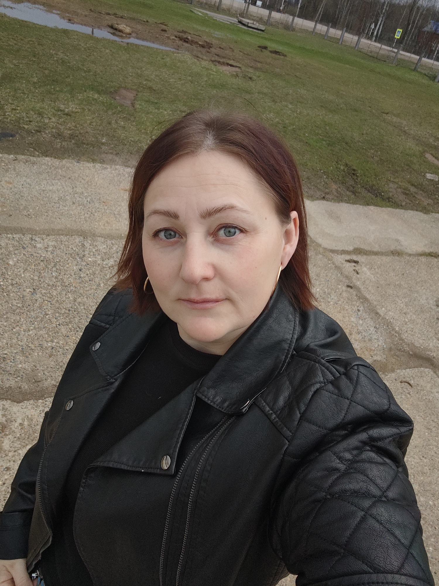 A woman with fair skin, light blue eyes, and reddish-brown hair gazes directly at the camera with a neutral expression. She appears to be in her 30s or 40s. She is wearing a black, possibly faux leather, motorcycle-style jacket featuring quilted patterns on the shoulders and upper sleeves, over a dark top. She accessorizes with small gold hoop earrings.
The woman is taking a selfie or being photographed from a slightly elevated, downward-angled perspective. The setting is outdoors in what appears to be Dubovaya, Russia. In the immediate foreground, she stands on a textured concrete or gravel surface. Behind her, a grassy area stretches into the midground, interspersed with patches of bare earth and possibly puddles, suggesting damp conditions or recent rain. The trees in the background are bare, indicating either early spring or late autumn. A fence runs along the treeline, and further in the distance, a yellow square road sign with an indecipherable black symbol is visible on the right. The lighting is soft and diffused, characteristic of an overcast day or late afternoon. There is no visible text in the image.