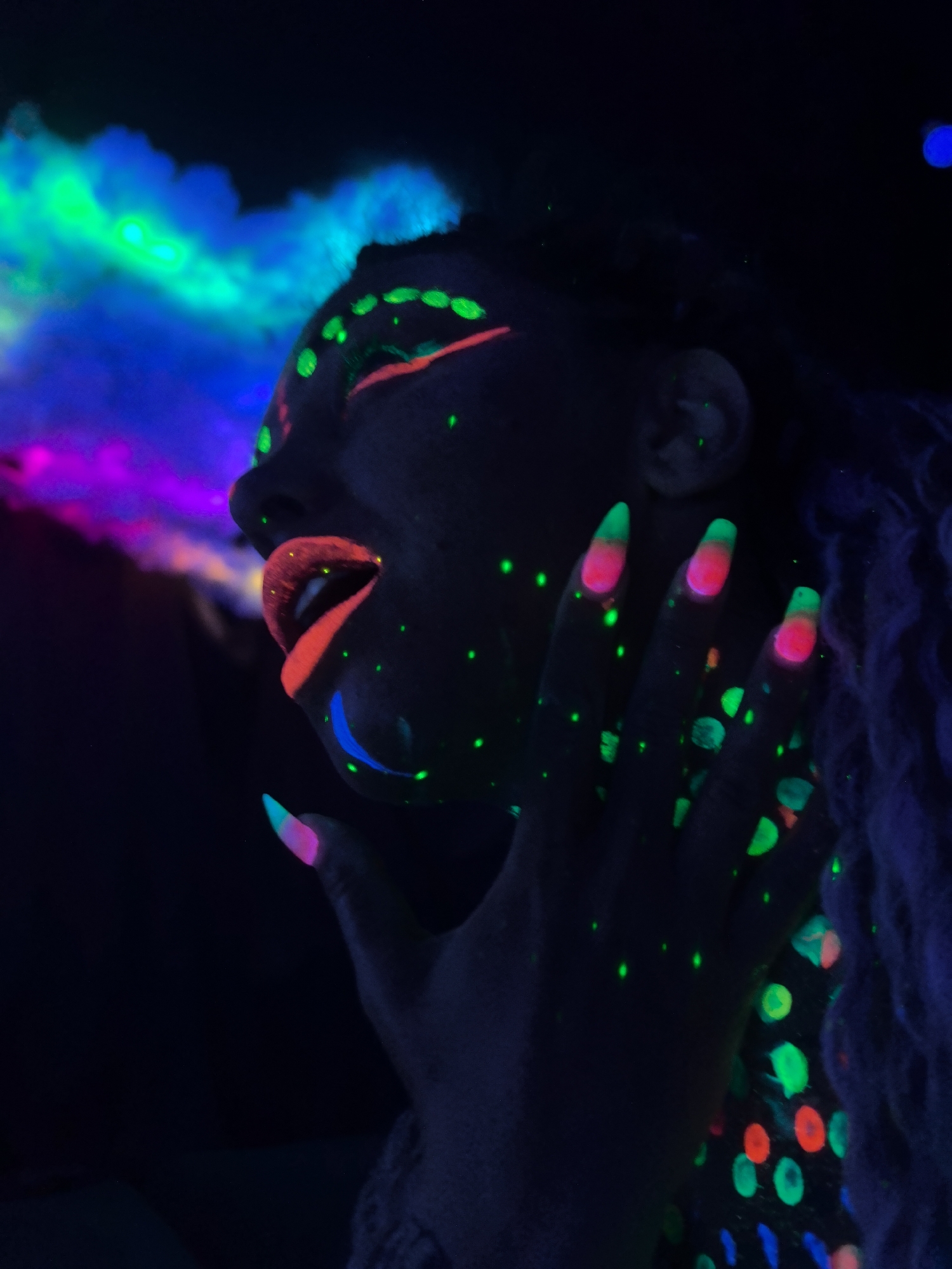 A profile view captures a person with dark skin under blacklight illumination. The subject's face is decorated with vibrant fluorescent makeup, highlighting the lips in red, the jawline in blue, and the eyelids in orange. Small green dots are scattered across their face and hand. The person's hand is raised near their face, with multi-colored nails glowing brightly. Fluorescent vapors in shades of blue, green, and purple emanate from the vicinity of their head. The environment is dark, contrasting with the colorful glow of the makeup and vapor. The location is not discernible from the image.