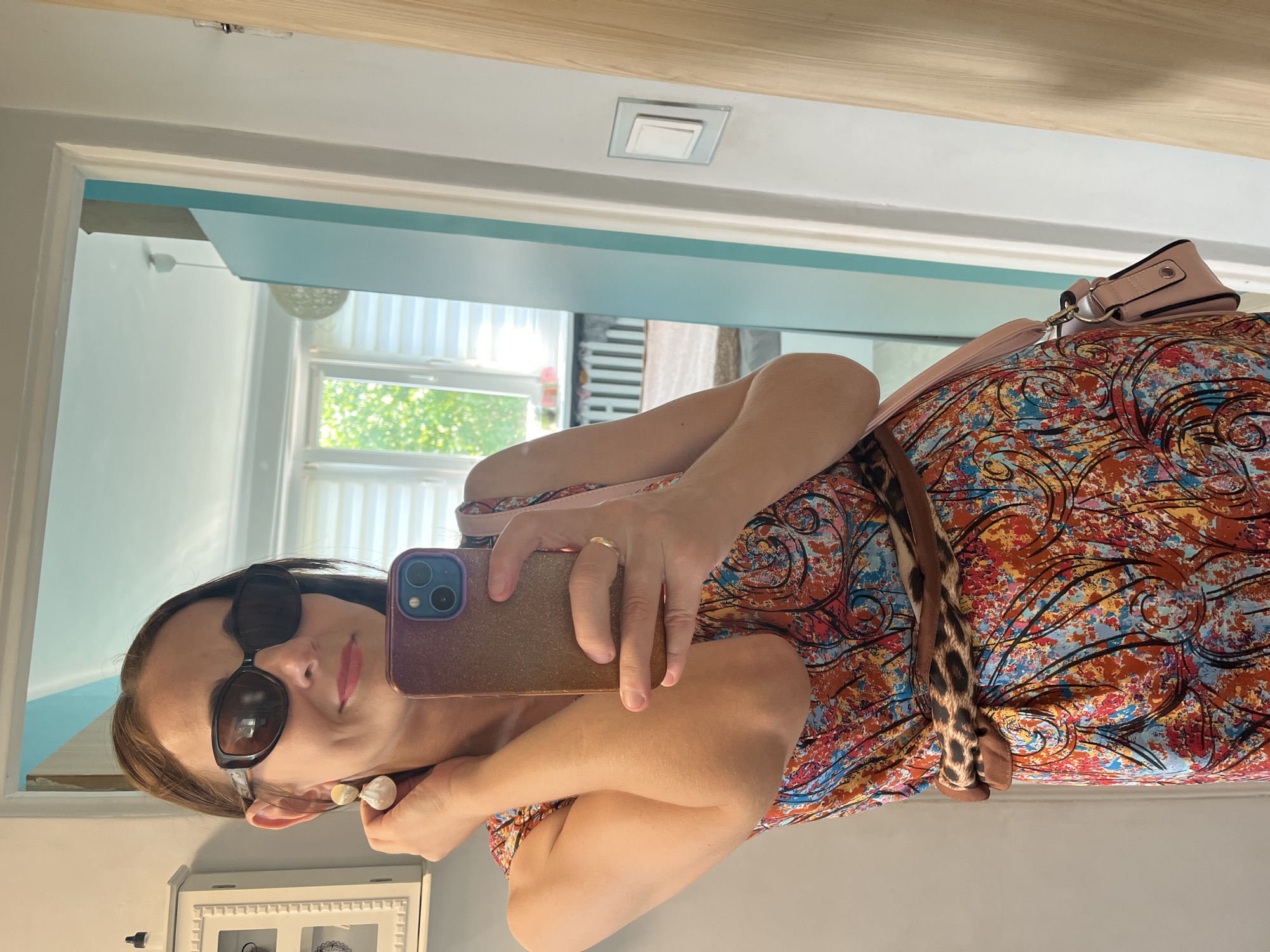 A woman is taking a mirror selfie in an interior location. She is wearing a colorful, patterned dress, a leopard print belt, and large sunglasses. She has on large, dangling earrings and a ring on her finger. A pink crossbody bag is slung over her shoulder. Her hair is dark brown and pulled back. The background appears to be a home interior, with a doorway, wall, and window visible.