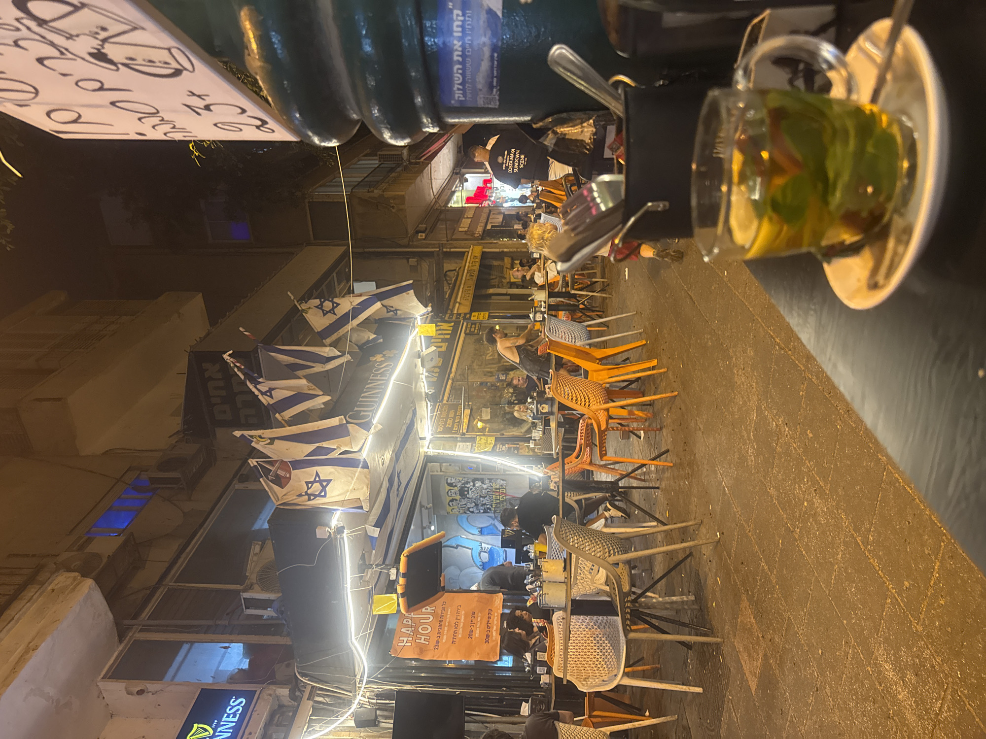 The image depicts a street-level view of an outdoor seating area for a restaurant or bar in Ramat Gan, Israel. Israeli flags are displayed, suggesting a patriotic or celebratory atmosphere. The scene is illuminated by string lights and ambient light from the establishment. Several patrons are seated at tables, engaged in conversation and dining. A sign promoting "Happy Hour" is visible, indicating the time of day. The foreground features a table with a glass of tea, complete with lemon and mint. In the background, a street with shops and buildings stretches into the distance. The overall impression is that of a lively evening scene at an establishment in a public urban space.