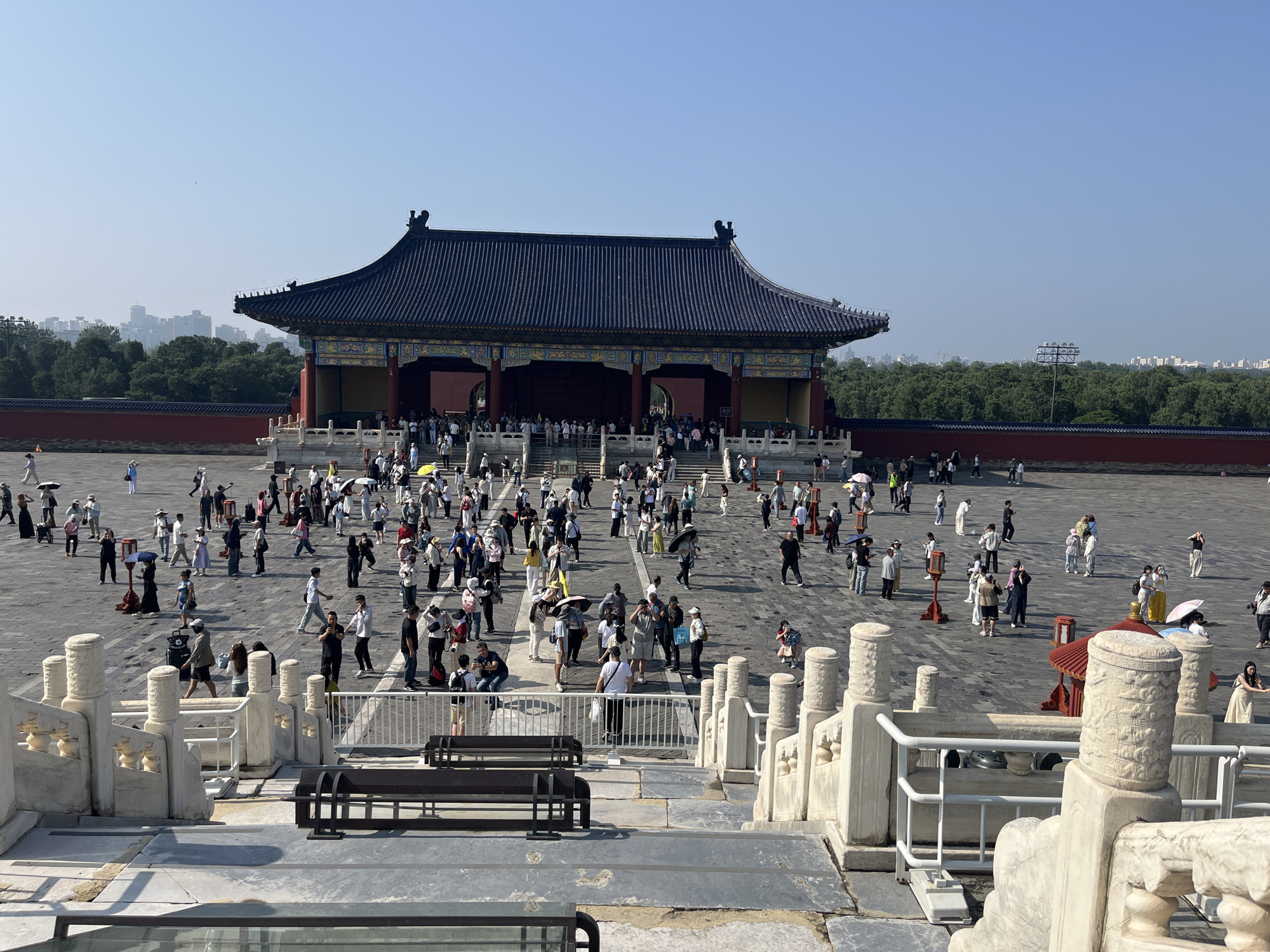 A large number of tourists are gathered in an outdoor courtyard in front of a traditional Chinese building with a dark blue tiled roof and ornate yellow and blue trim. The building has a wide entrance with multiple archways and is supported by red columns. The ground is paved with stone tiles, and there are stone balustrades and benches in the foreground. Many people are walking around, some are taking pictures, and some are holding umbrellas. The sky is clear and blue, and trees are visible in the background, with a cityscape in the far distance. The scene appears to be a tourist attraction in a city.