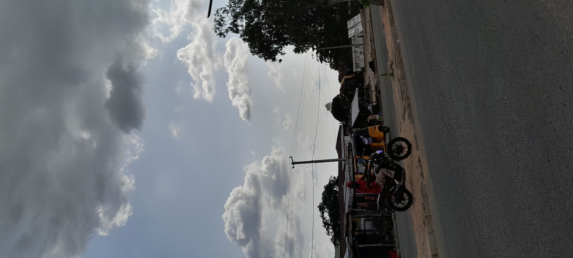 The image depicts a street scene in Jalingo, Nigeria, under a cloudy sky. In the foreground, a black motorcycle is parked on the side of the asphalt road. The rider, wearing a red shirt and camouflage-patterned pants, is seated on the motorcycle. Behind the motorcycle, a yellow auto-rickshaw, also known as a Keke Napep, is visible, with passengers inside. The background features a row of informal shops or stalls with corrugated metal roofs, and some trees. A utility pole with overhead wires is positioned in the middle ground. The overall environment suggests a typical urban or semi-urban setting in Nigeria.