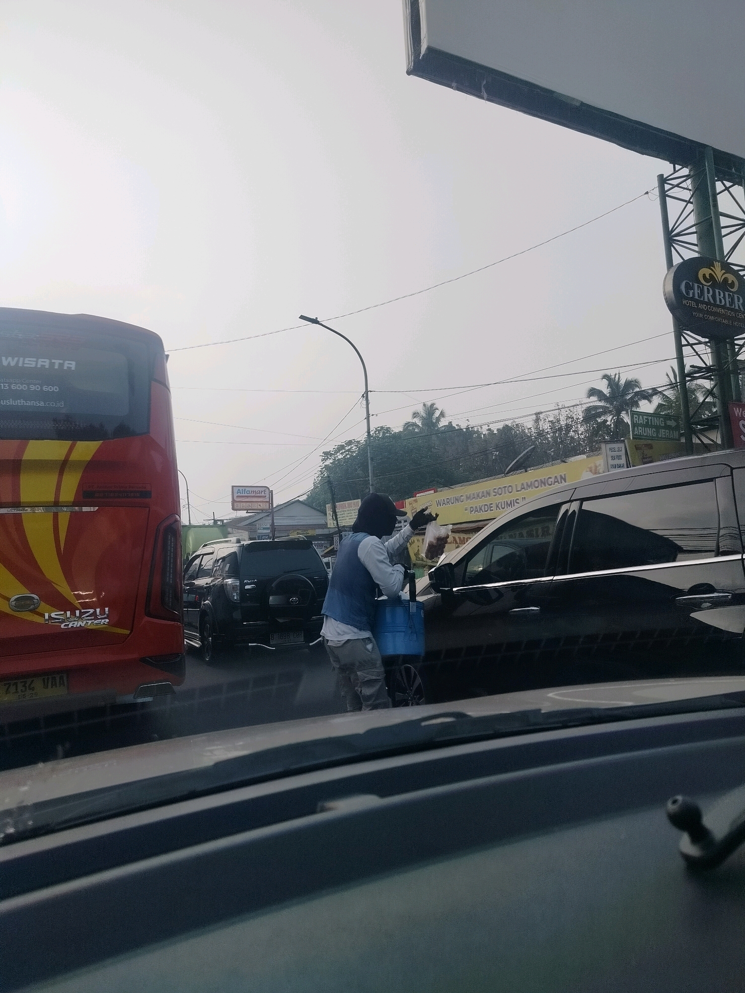 The image depicts a street scene in Megamendung, Indonesia, characterized by moderate traffic. In the foreground, the interior of a car is visible, including the dashboard and windshield. To the left, the rear of a red bus with "WISATA" and "ISUZU CANTER" branding is parked. To the right of the bus, a person wearing a blue vest over a white shirt and dark pants is seen interacting with a dark-colored SUV. The person is holding a plastic bag and a blue bucket. In the background, a yellow sign for "WARUNG MAKAN SOTO LAMONGAN 'PAHDE KUMIS'" is visible, along with other signs for "Alfmart" and "RAFTING ARUNG JERAM". Palm trees and utility poles are also present in the background, suggesting an outdoor, possibly humid environment. The sky is overcast and bright, indicating daylight.