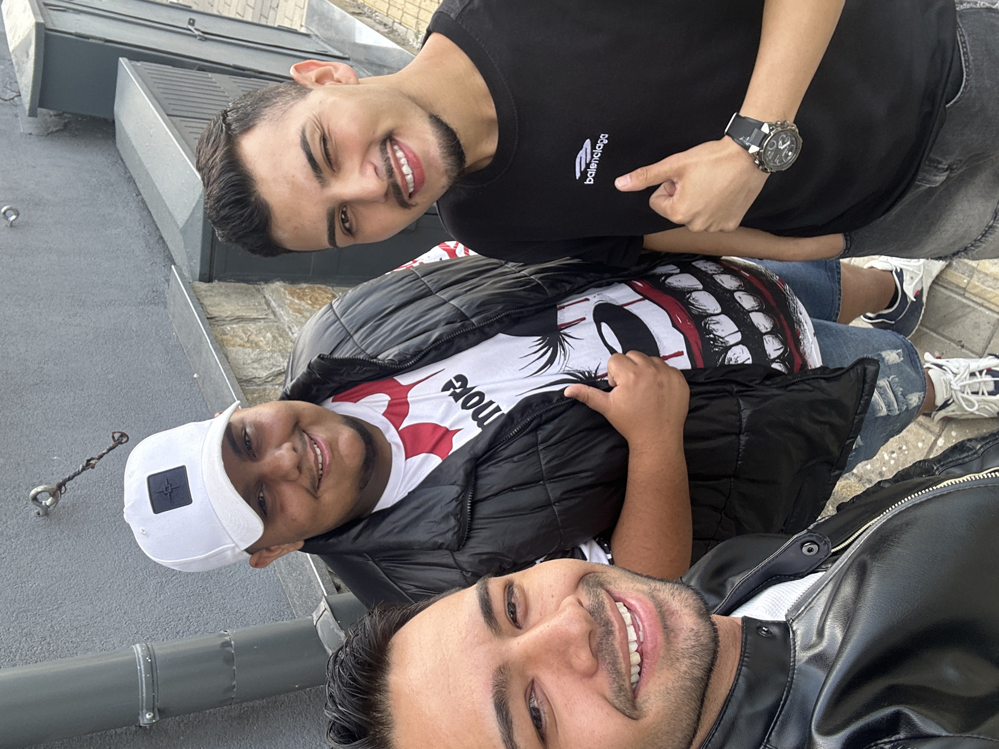 A close-up outdoor photograph depicts three young adult males standing together in an urban environment in Sofia, Bulgaria, during daylight.
On the left, partially cropped, a male with dark hair and a light beard smiles directly at the camera. He wears a black leather jacket over a white garment, visible at the collar.
In the center, a male of heavier build smiles, wearing a white baseball cap with a dark square emblem (identifiable as a Stone Island badge), a black puffy vest over a white t-shirt featuring a stylized cartoon mouth graphic with prominent red accents and the word "more," and ripped blue denim shorts. His right thumb is raised in a 'thumbs-up' gesture. White sneakers with blue accents are visible on his feet.
On the right, a third male winks with his right eye and smiles, displaying a short beard and mustache. He is dressed in a black short-sleeved t-shirt with a white "balenciaga" logo printed on the chest, and dark grey ripped denim shorts. A large, dark-faced wristwatch with a black band is worn on his left wrist. His left thumb is also raised in a 'thumbs-up' gesture.
All three individuals convey a friendly demeanor through their smiles and positive hand gestures.
The background consists of a textured grey building wall. A grey metal downspout runs vertically on the left. Above the central figure, a metal hook with a twisted dark object, possibly an old wire or strap, is attached to the wall. To the right, grey outdoor utility boxes are mounted on the wall. The ground is paved with light-colored square tiles. Below the grey wall, a section of light-colored stone masonry is visible. Natural, soft daylight illuminates the scene.